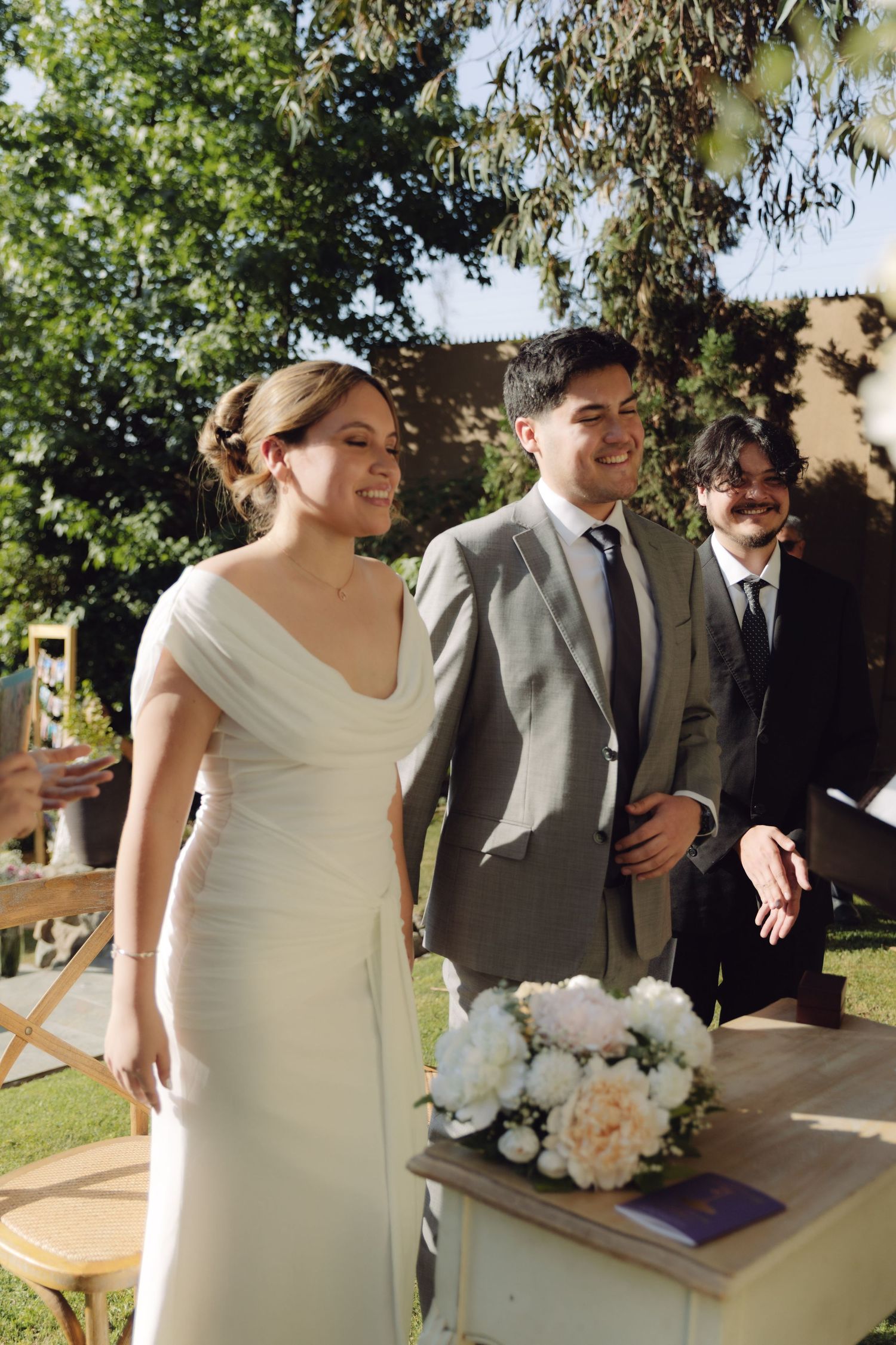 Happy bride and groom smiling together beside white peony bouquet at outdoor wedding.