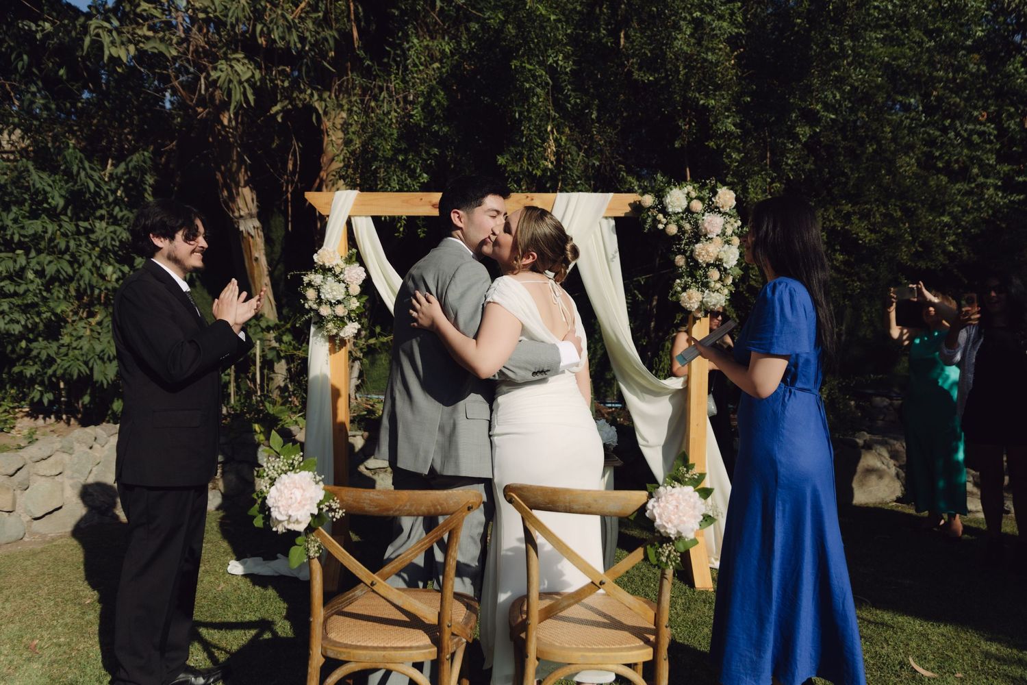 Bride and groom share first kiss under wooden floral arch at outdoor wedding ceremony.