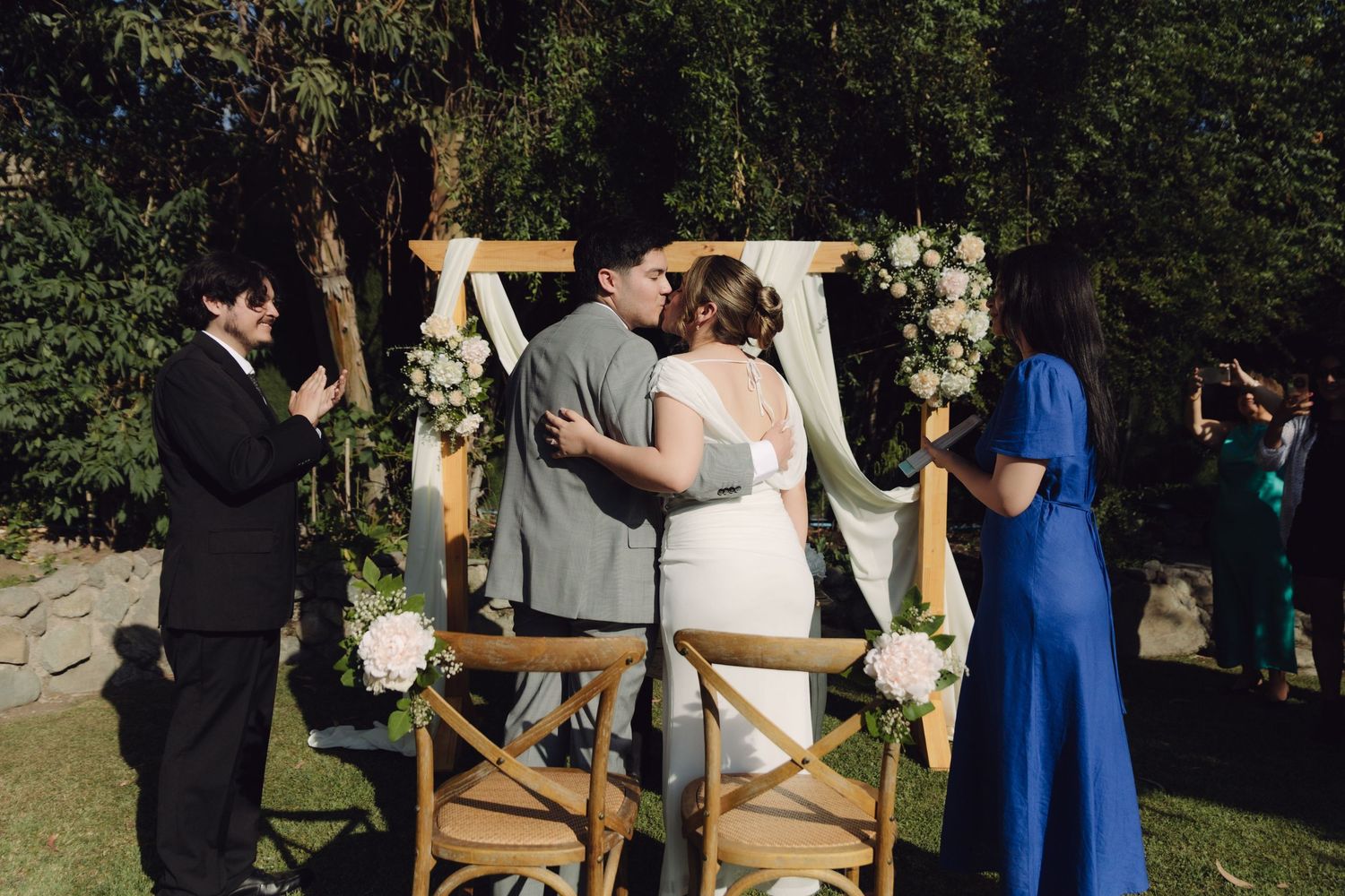 Newlyweds kiss under decorated wooden arch during outdoor garden wedding ceremony.