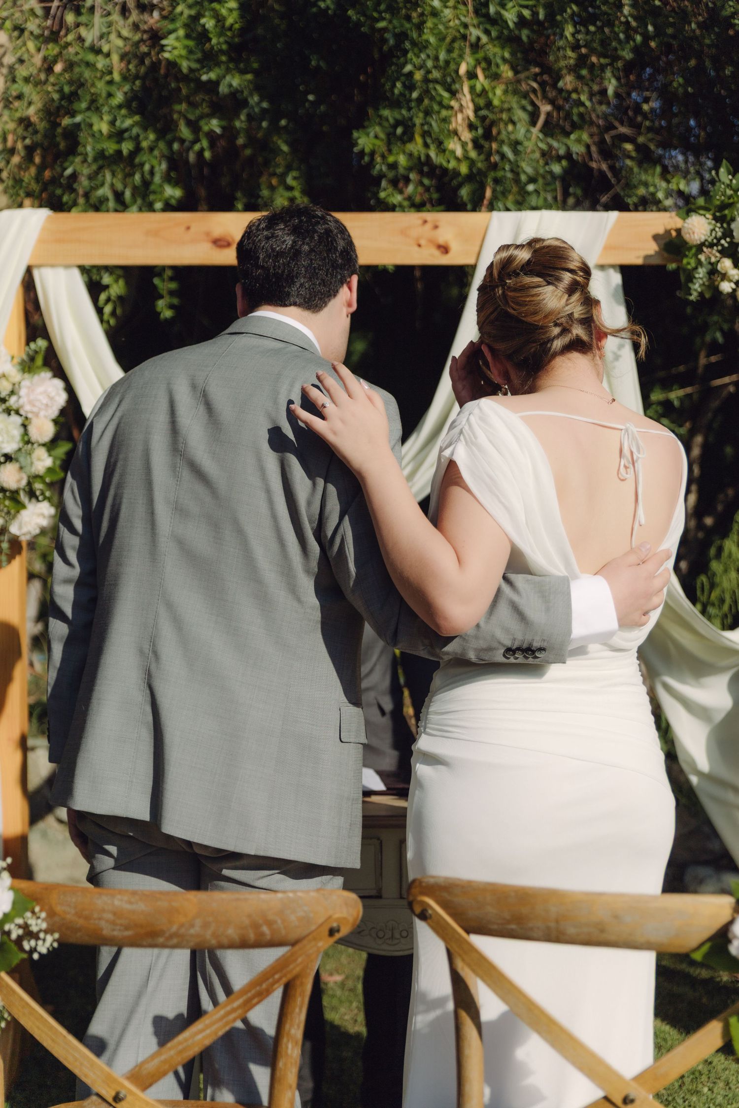 Bride and groom stand together at outdoor wedding altar decorated with wooden arch and white draping.
