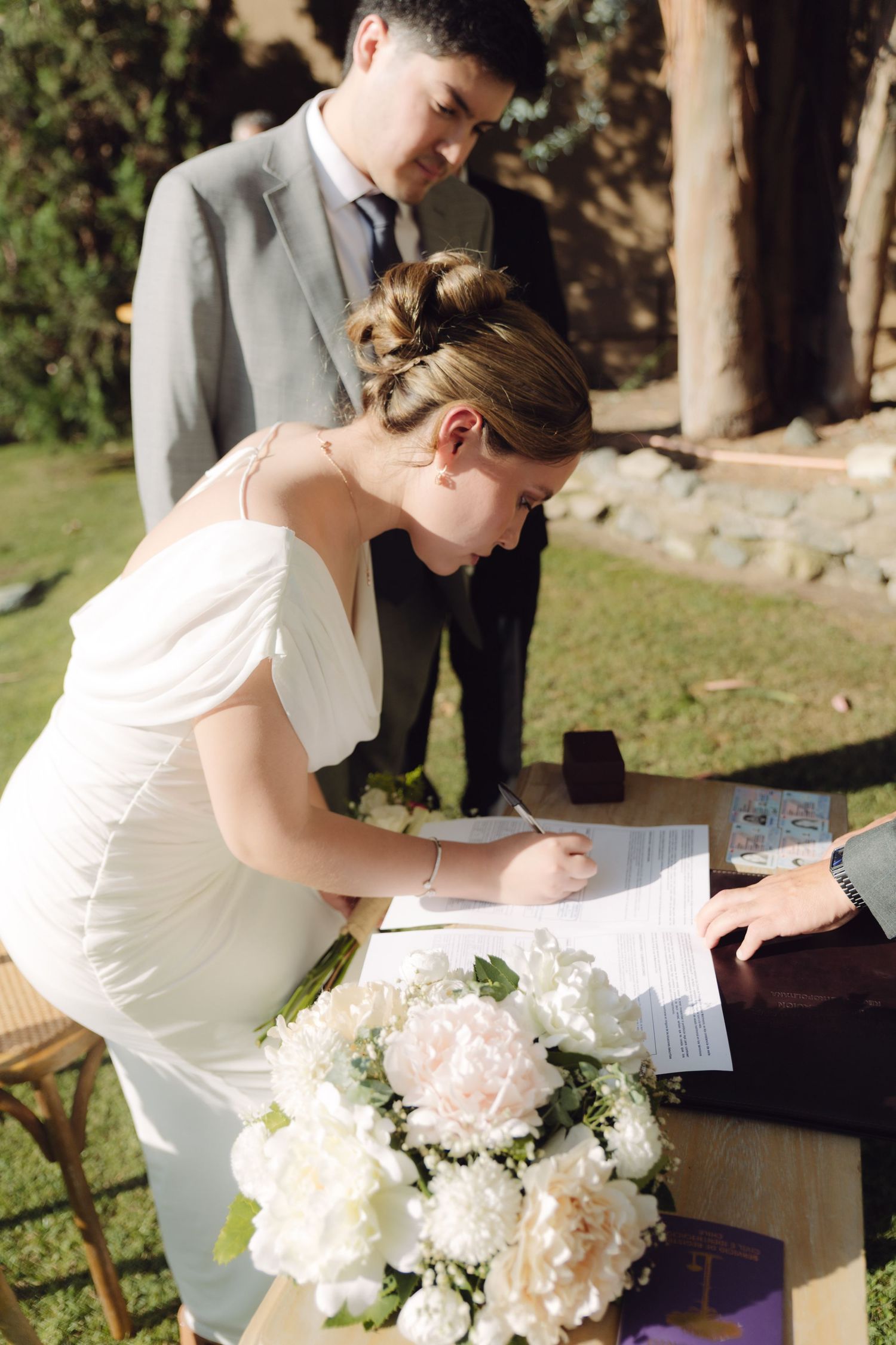 Bride in white dress signs wedding documents at outdoor ceremony with groom standing beside her.