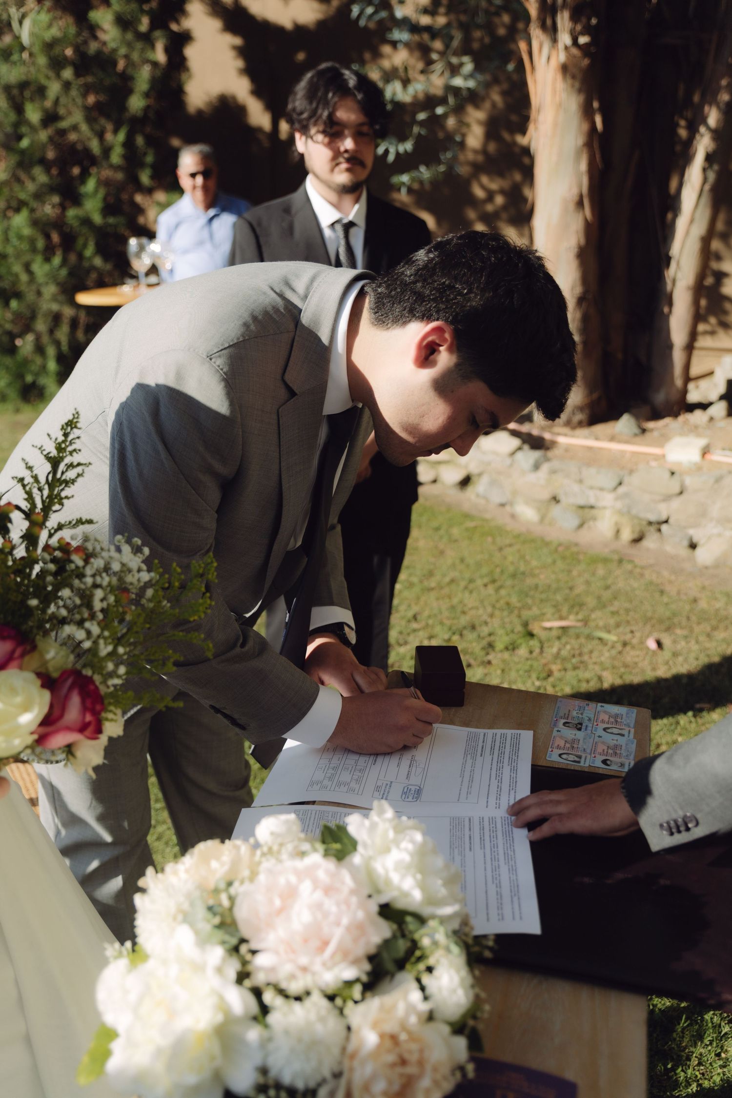 Groom in gray suit signs official wedding documents at outdoor ceremony altar with floral arrangements.