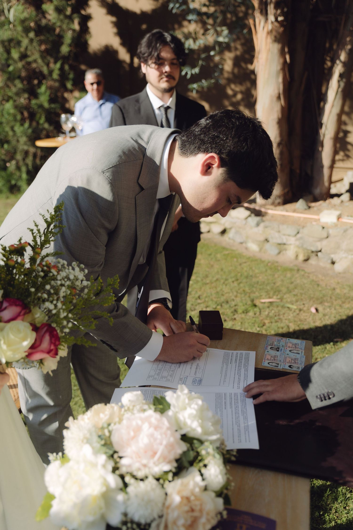 Groom in gray suit signs wedding documents at flower-adorned ceremony table outdoors.