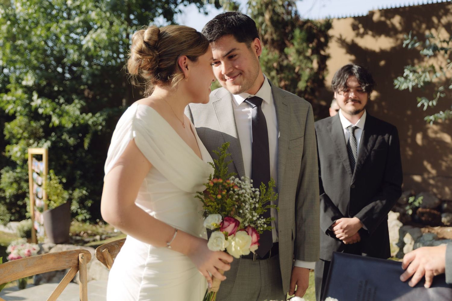 Smiling bride holding bouquet stands with groom at outdoor wedding ceremony.