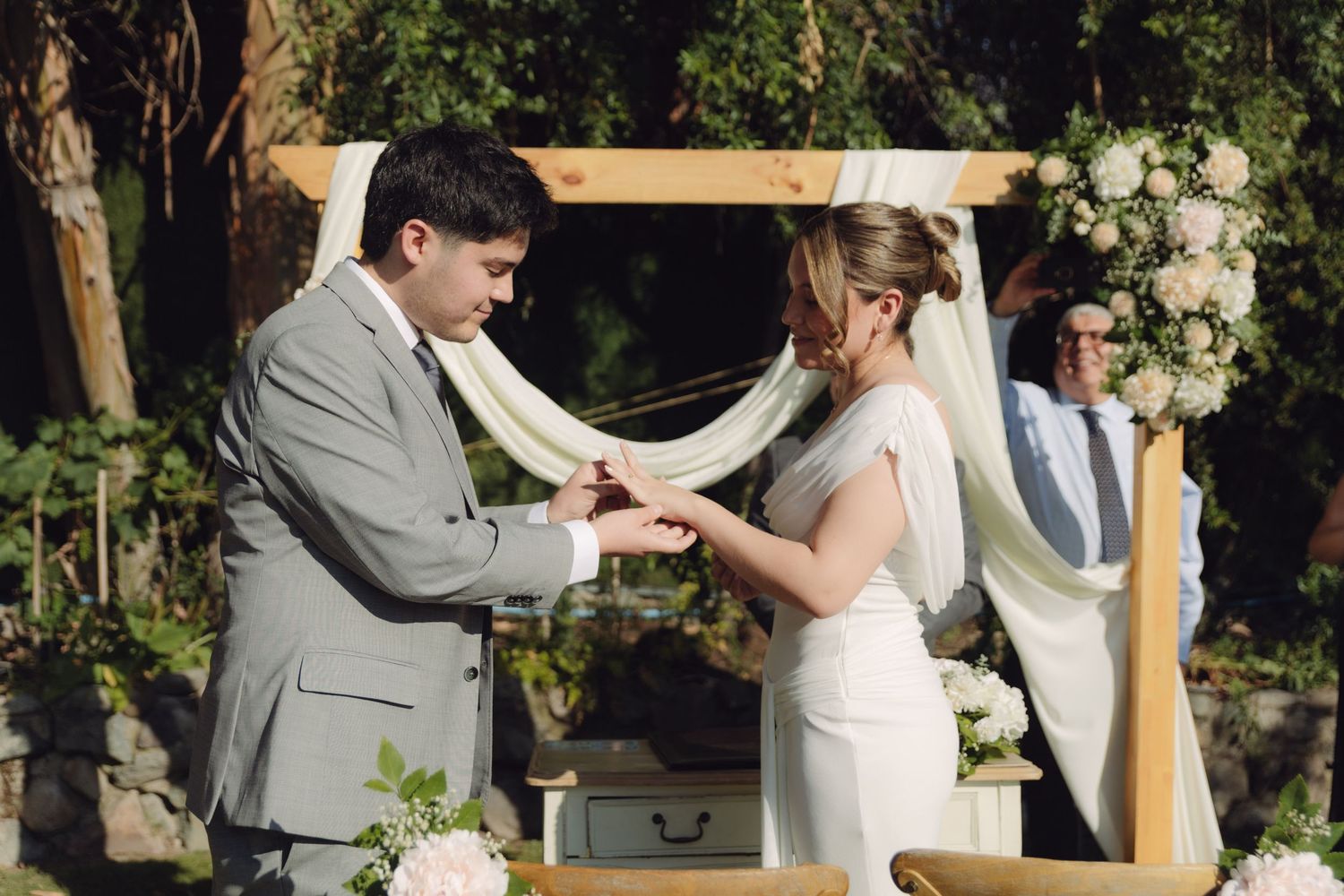 Bride and groom exchange rings under decorated wooden wedding arch with white draping and flowers.