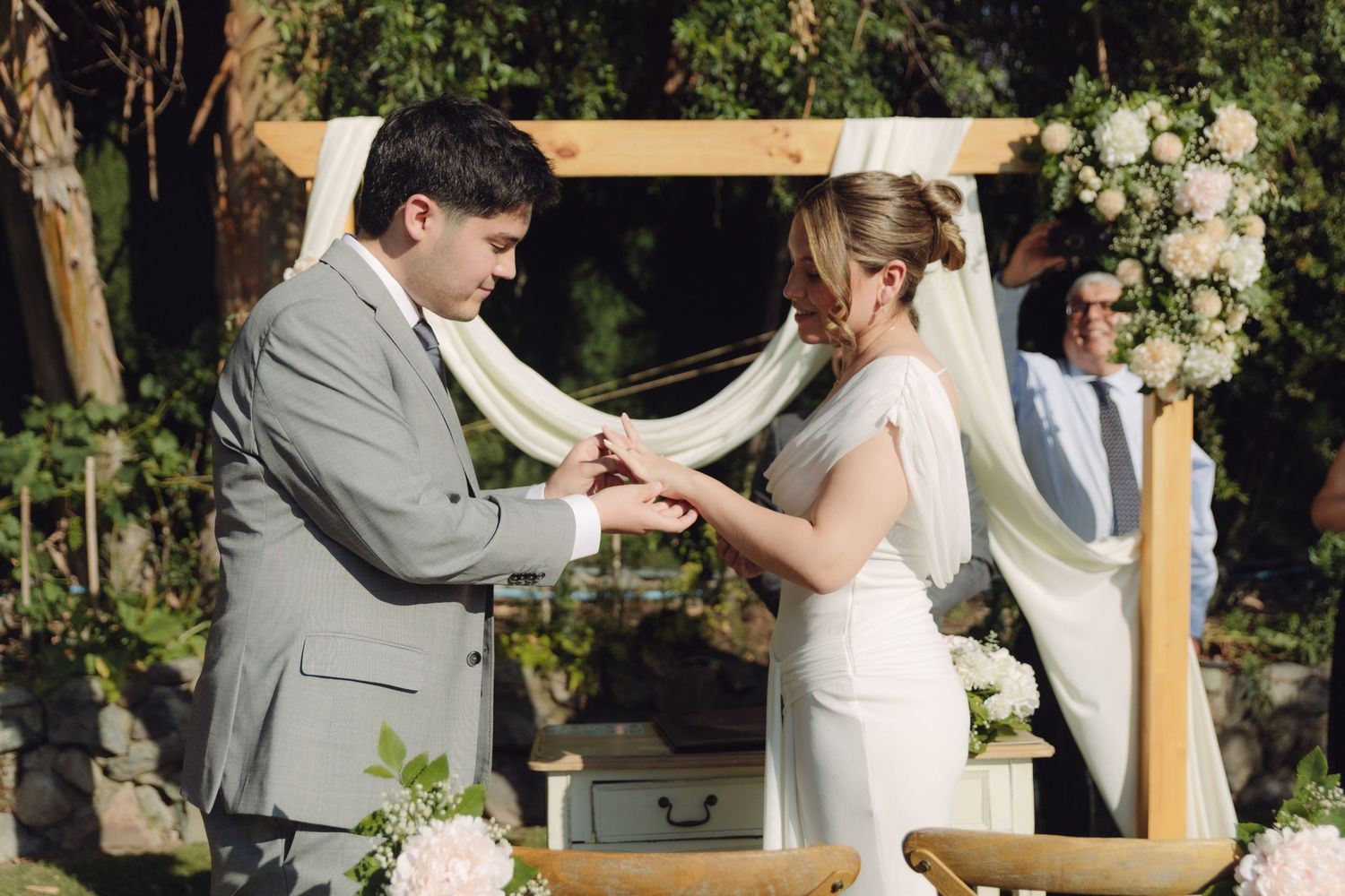 Groom places wedding ring on brides finger under floral wooden arch at outdoor ceremony.