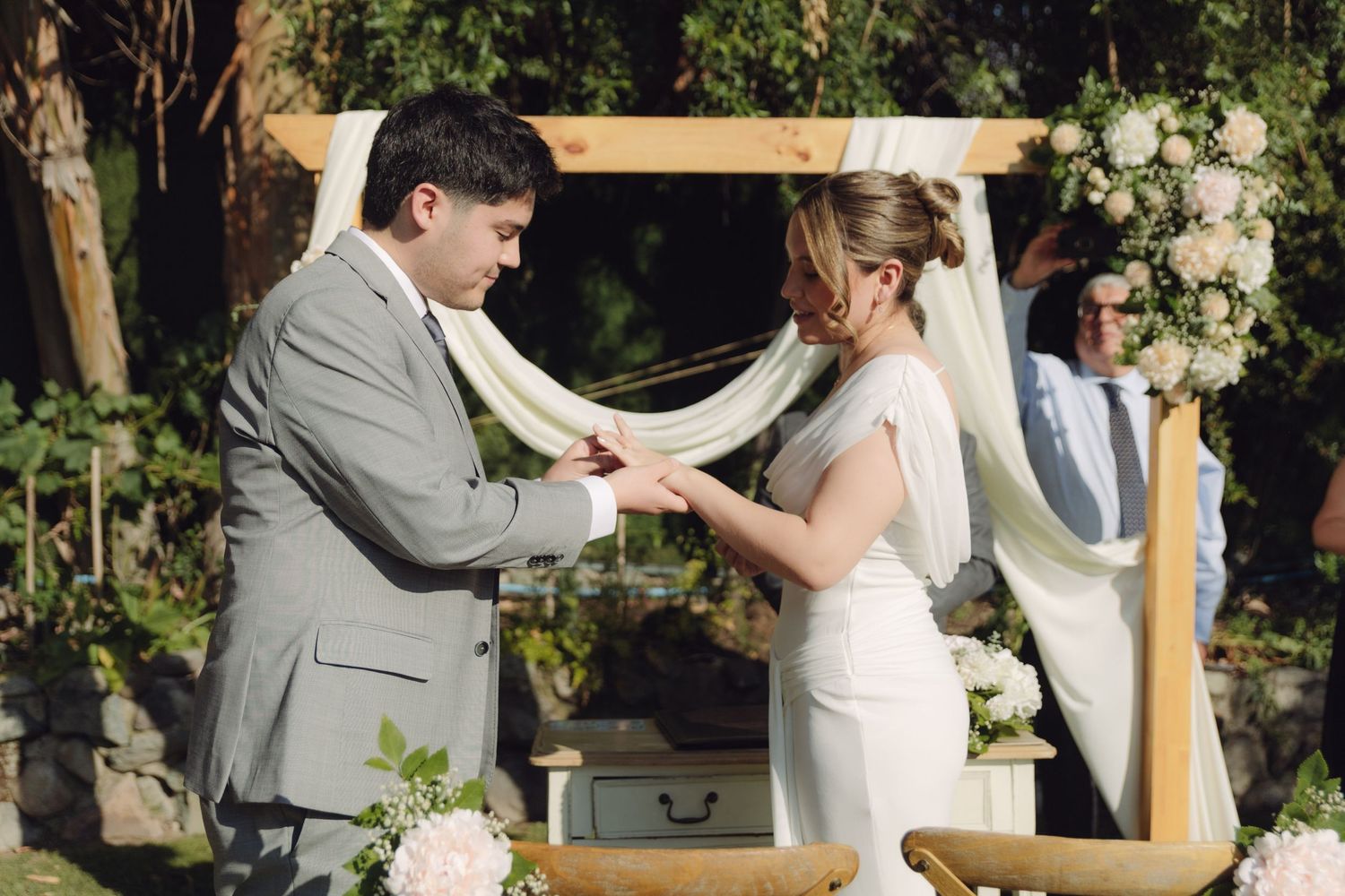 Groom places wedding ring on brides finger during outdoor ceremony under wooden floral arch.