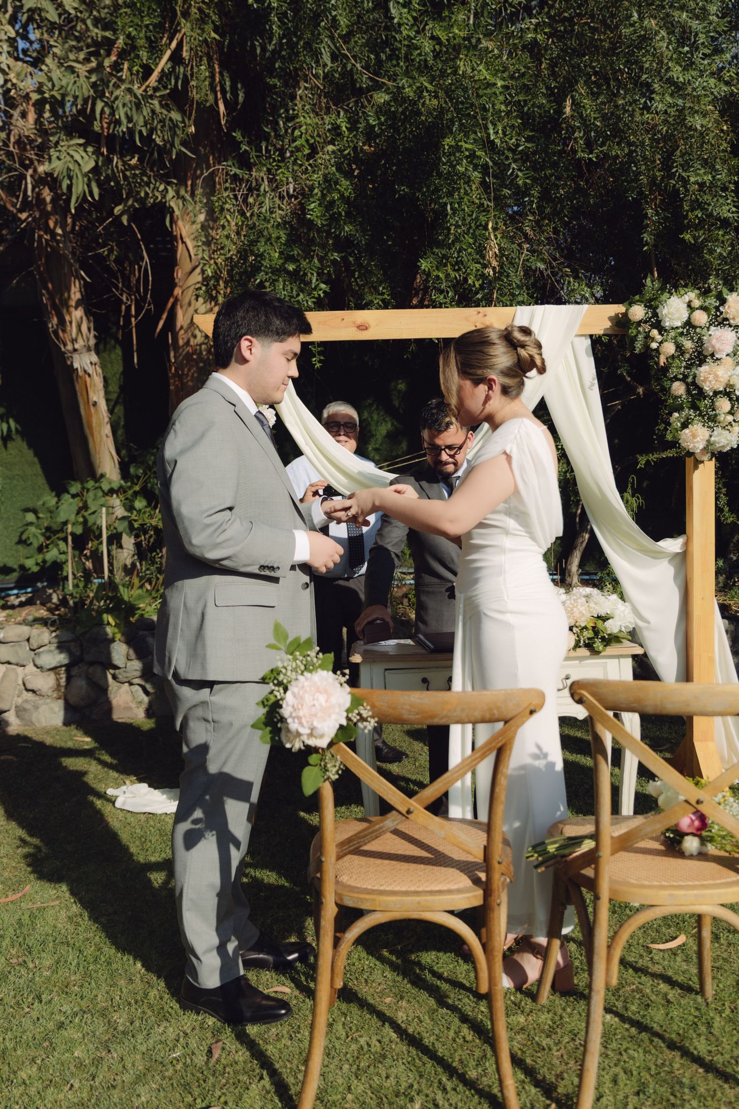 Bride and groom perform ring exchange ceremony outdoors at wooden arch altar with pink flowers.