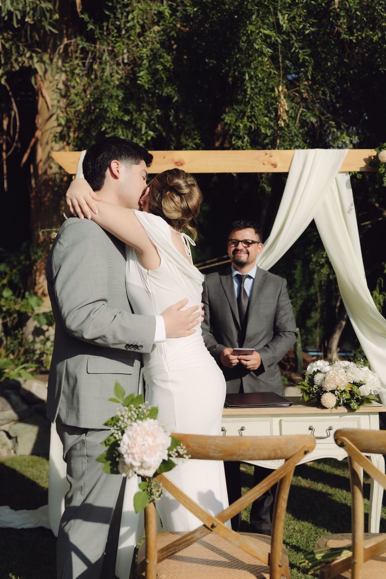 Bride and groom embrace after exchanging vows as officiant watches at outdoor ceremony.