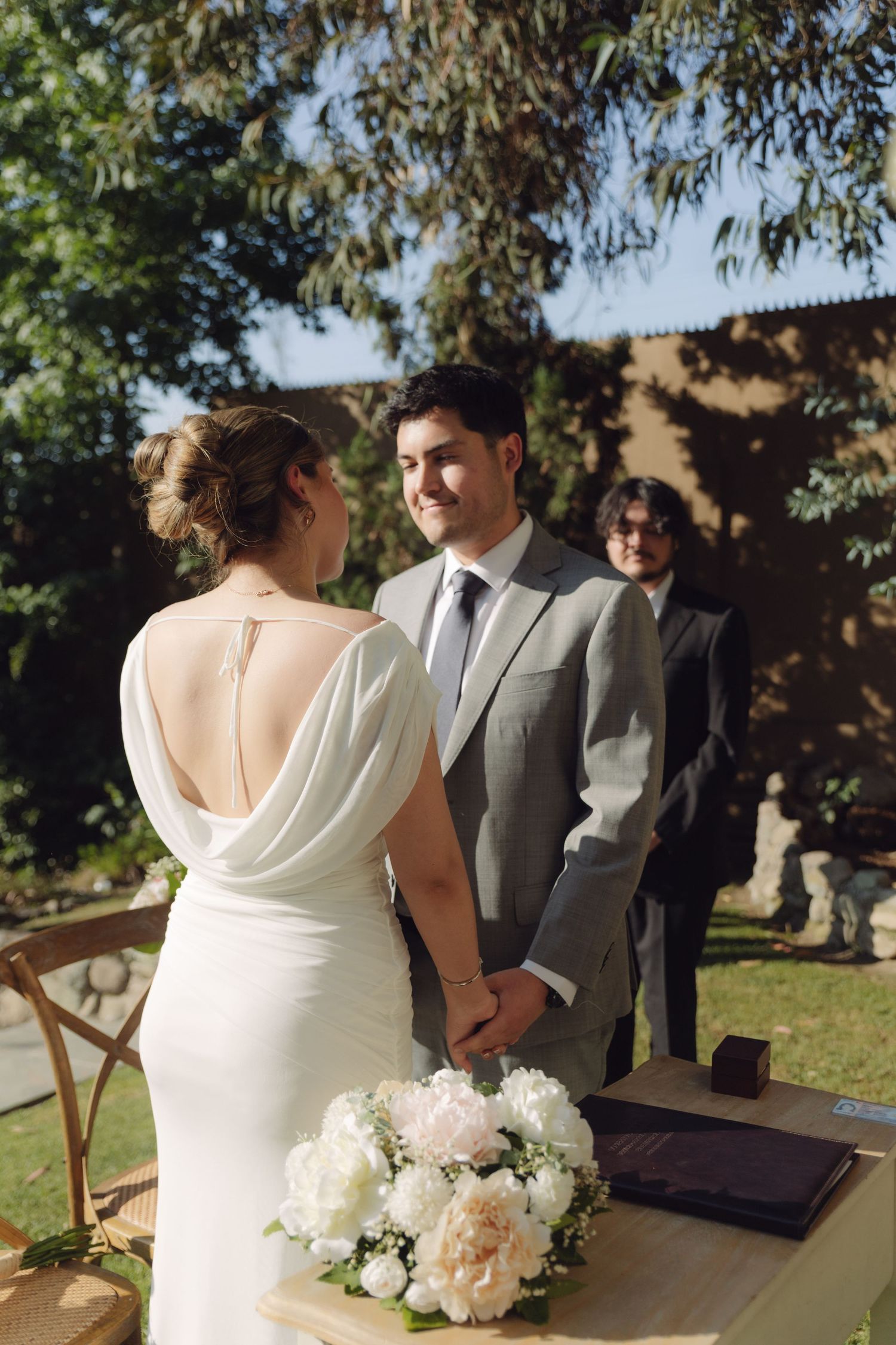Smiling groom holds brides hands at outdoor wedding ceremony with white peony bouquet on table.
