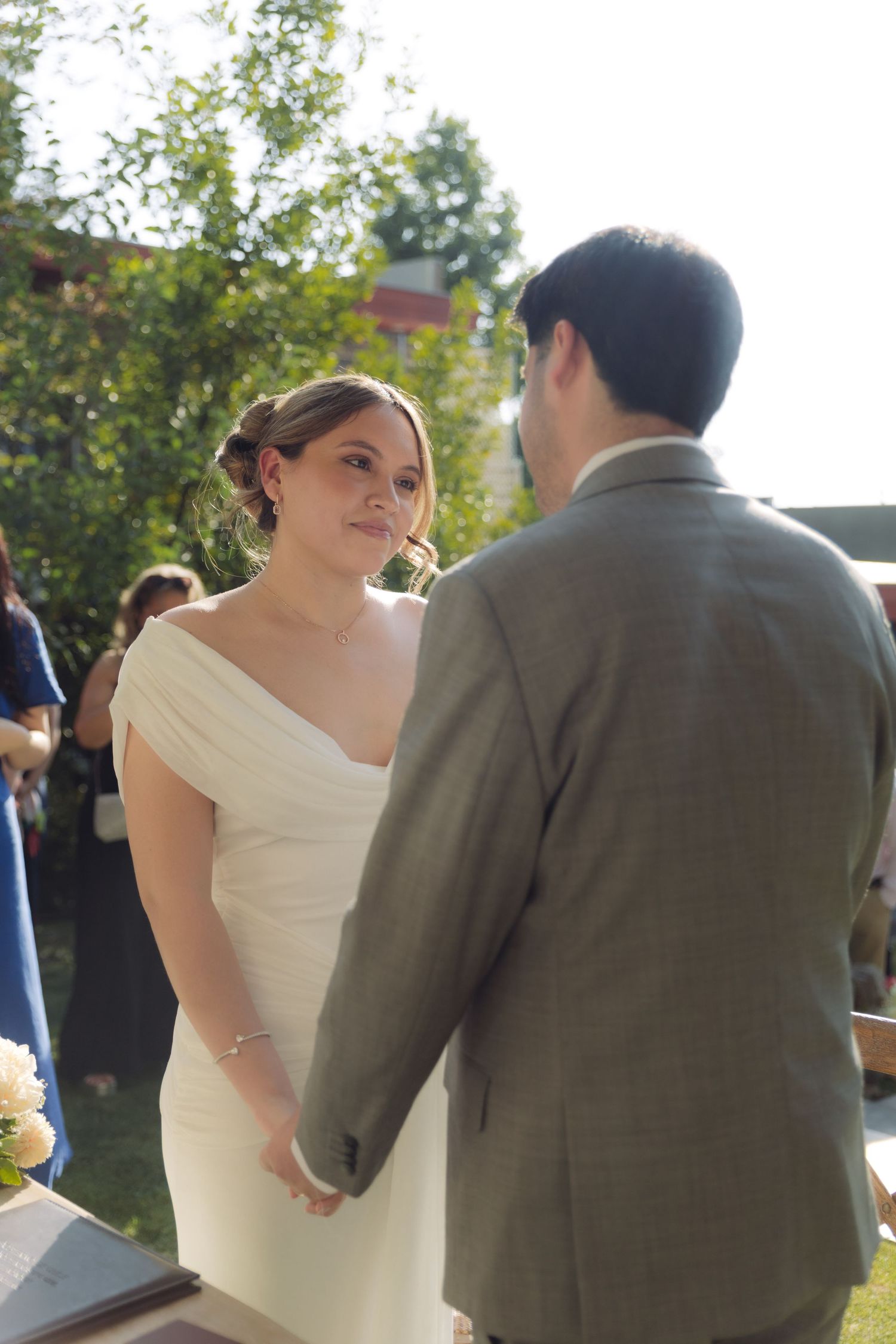 Bride in white off-shoulder gown holds hands with groom during outdoor wedding ceremony vows.