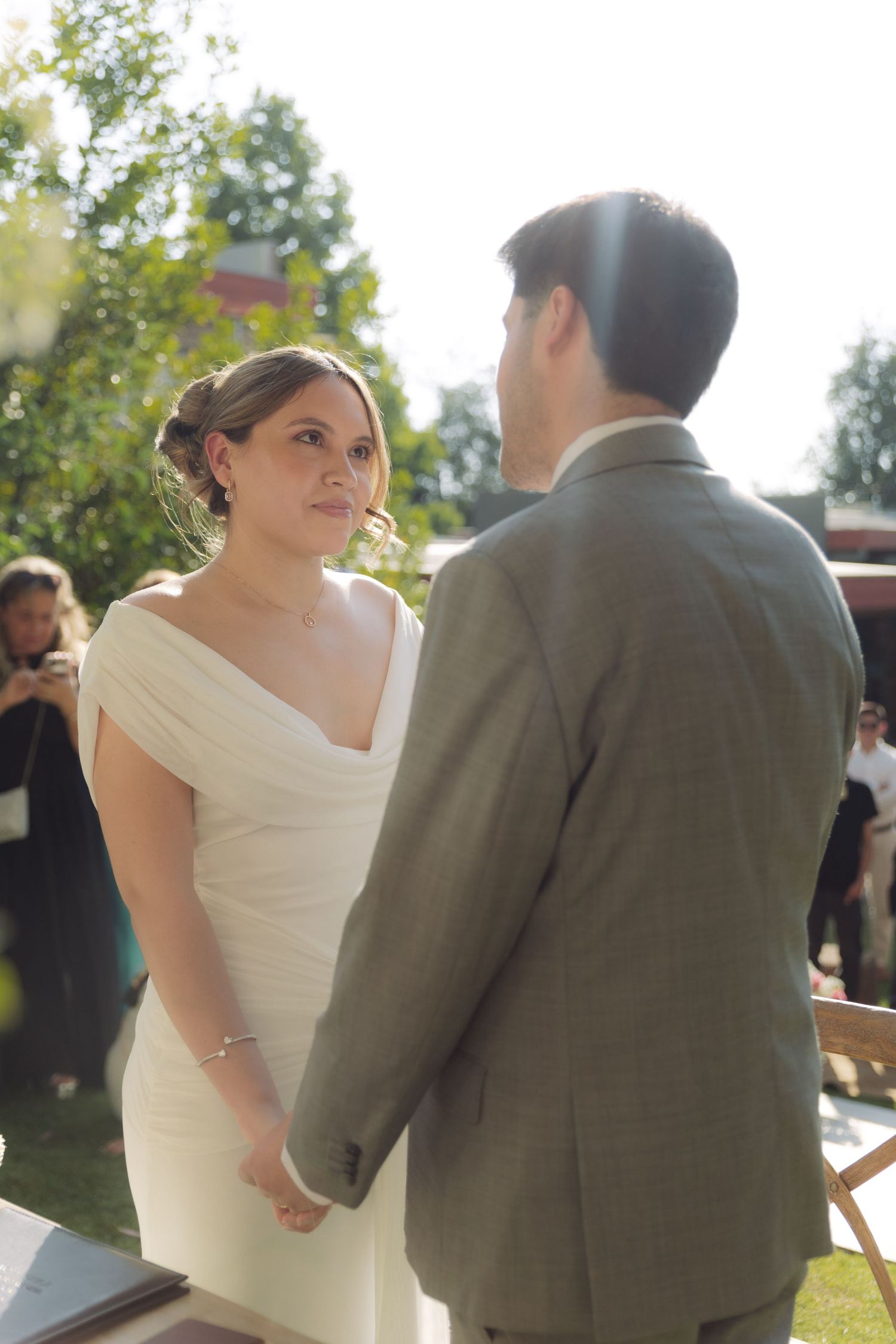 Bride gazes at groom while holding hands during sunlit outdoor wedding ceremony in garden.
