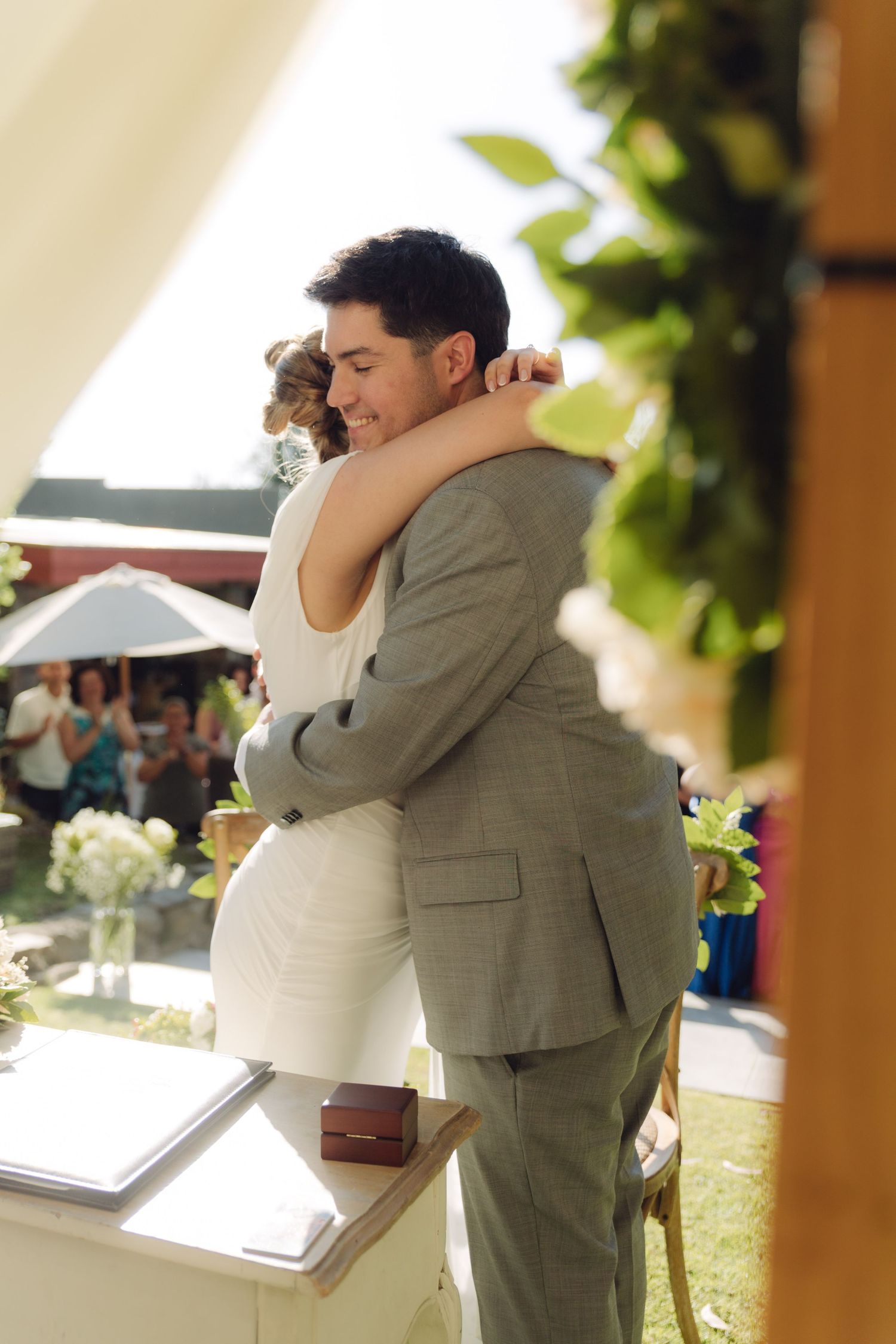 Newlyweds share joyful hug at outdoor wedding ceremony as guests celebrate in background.