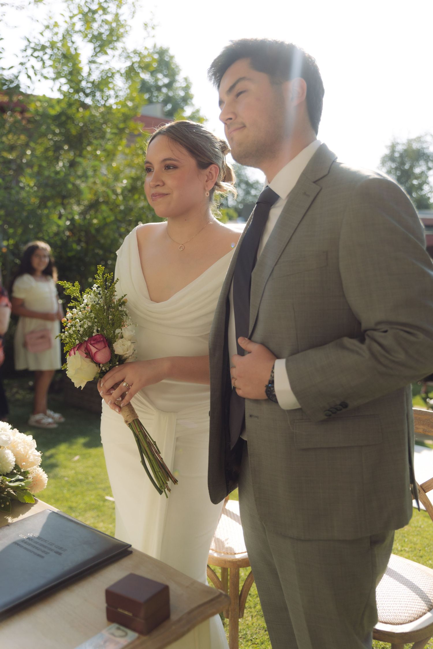 Bride holding bouquet and groom in gray suit stand together at outdoor wedding altar.