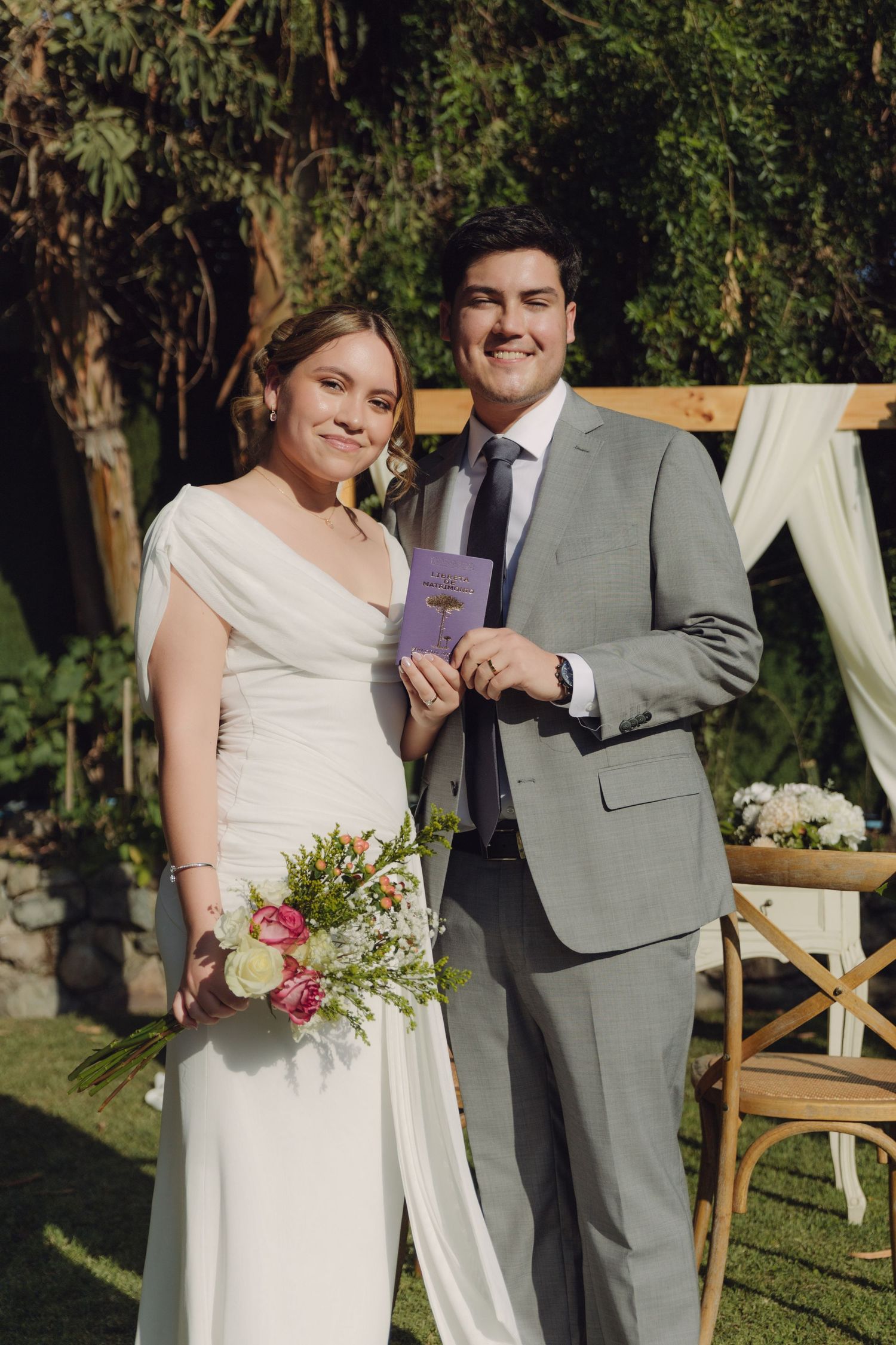 Smiling bride and groom posing outdoors at garden wedding ceremony with floral bouquet and wooden arch.