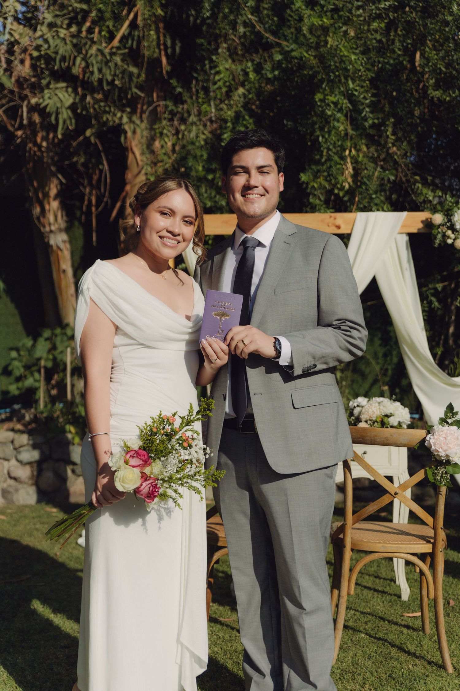 Smiling bride and groom pose together holding marriage certificate at outdoor wedding.