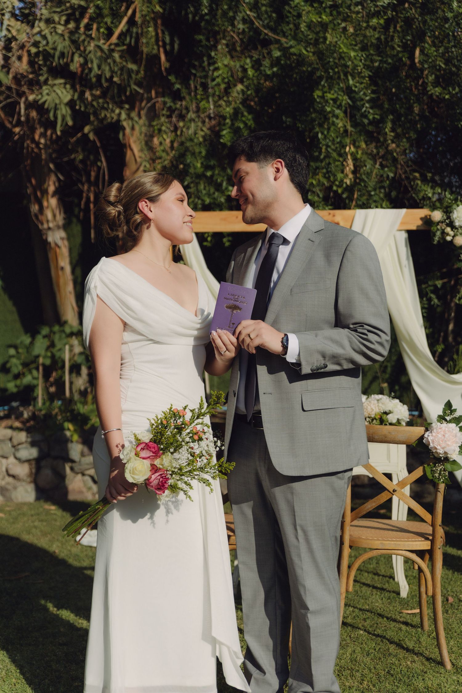 Bride and groom exchanging loving glances during outdoor garden wedding ceremony with wooden arch backdrop.