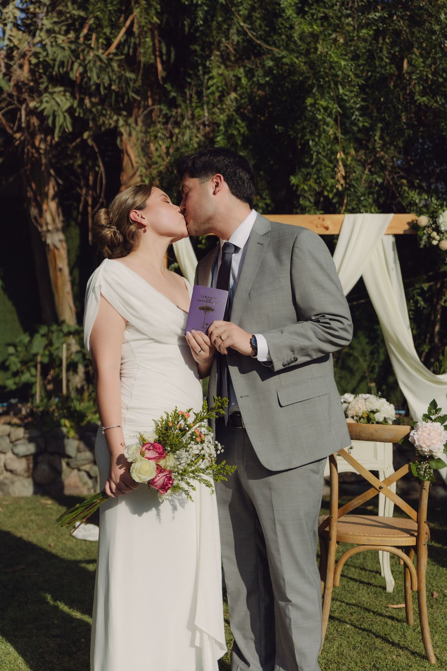 Bride and groom share a kiss while holding marriage certificate at garden wedding ceremony.