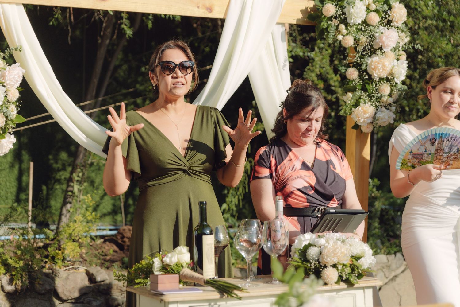Wedding officiant in green dress speaks at outdoor ceremony altar with floral decorations.