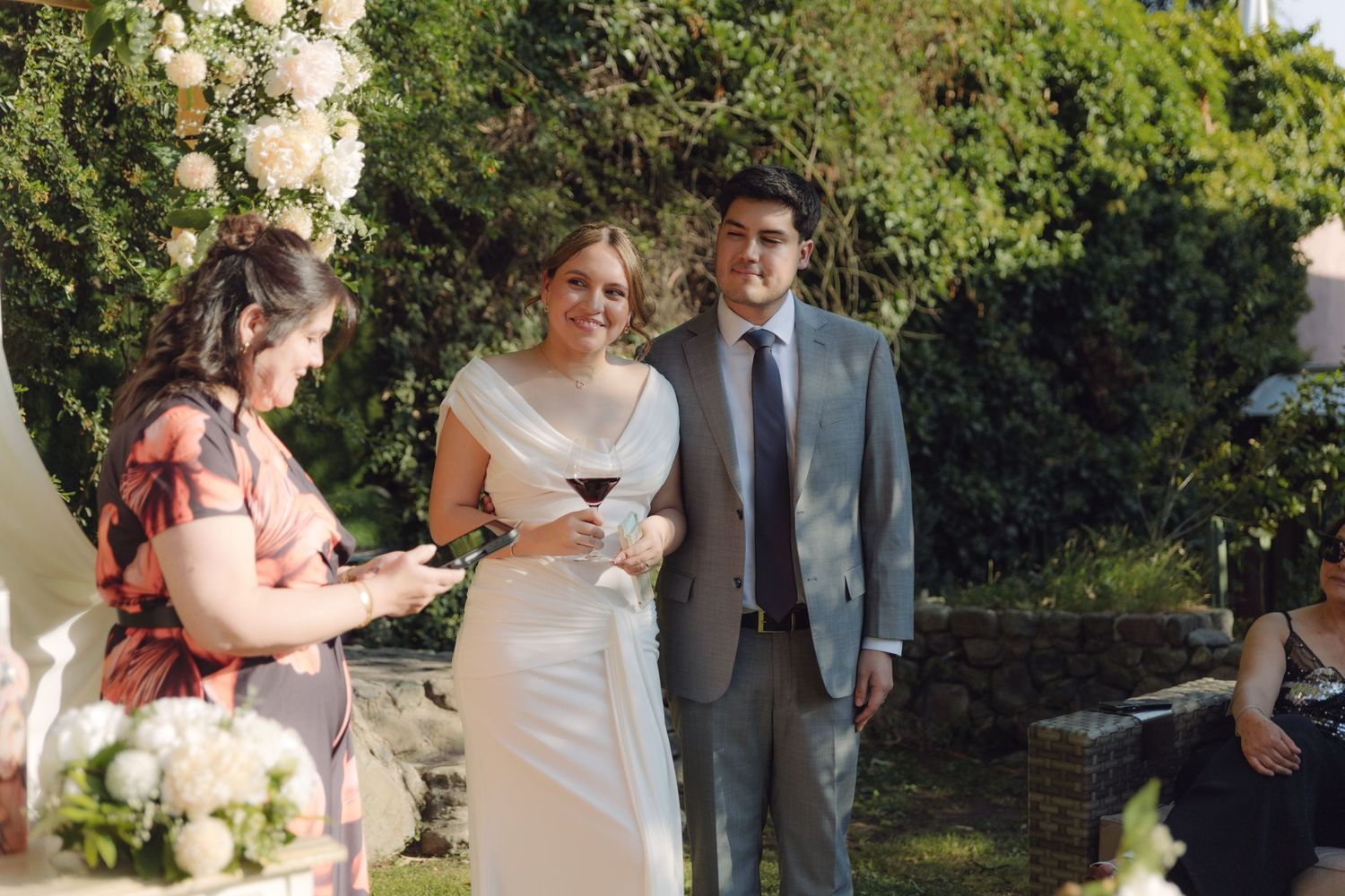 Bride holding wine glass smiles with groom during outdoor garden wedding ceremony.