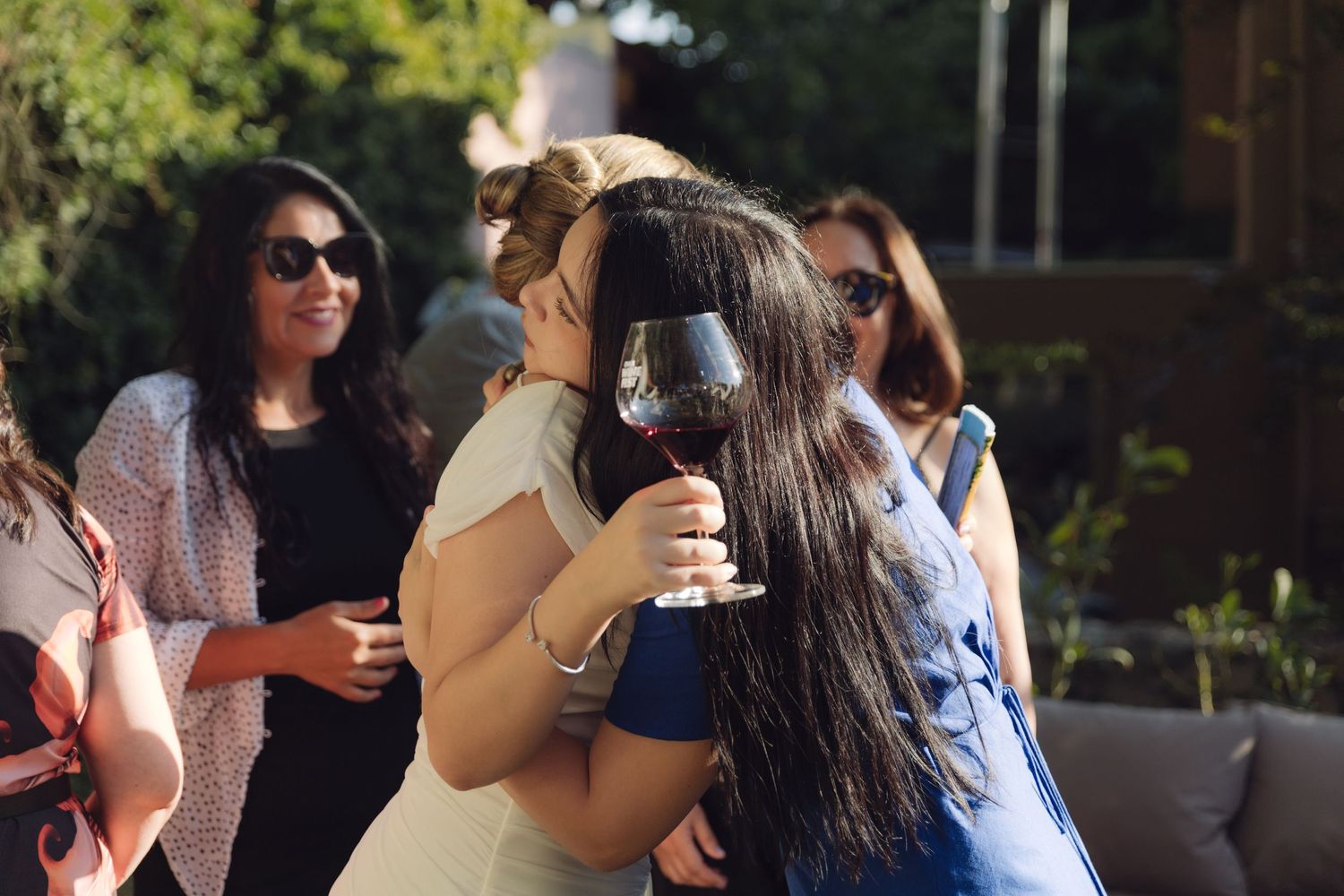 Bride hugging a dark-haired guest holding red wine at sunny outdoor wedding reception.