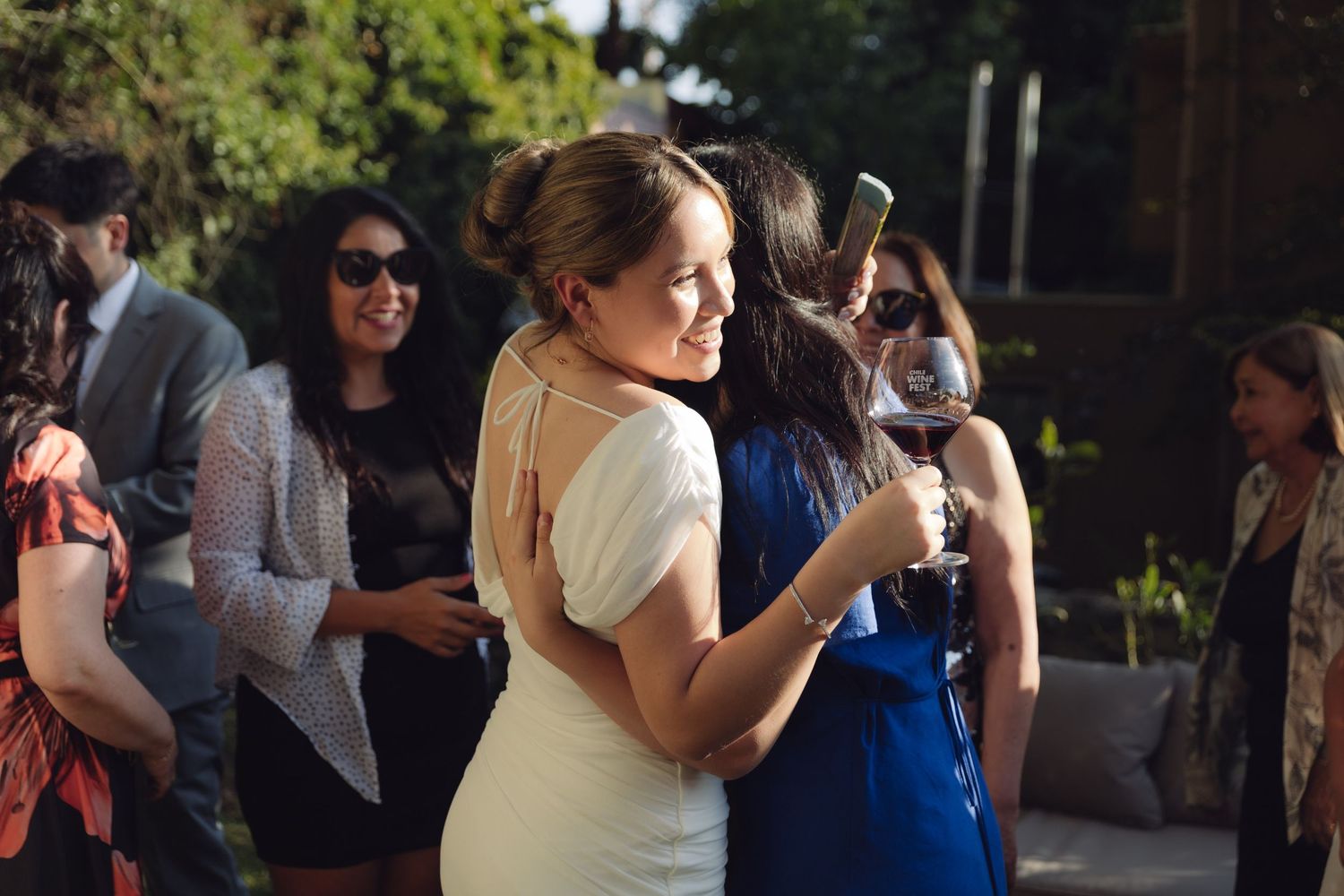 Smiling bride in white dress embraces a guest in blue at outdoor wedding reception.