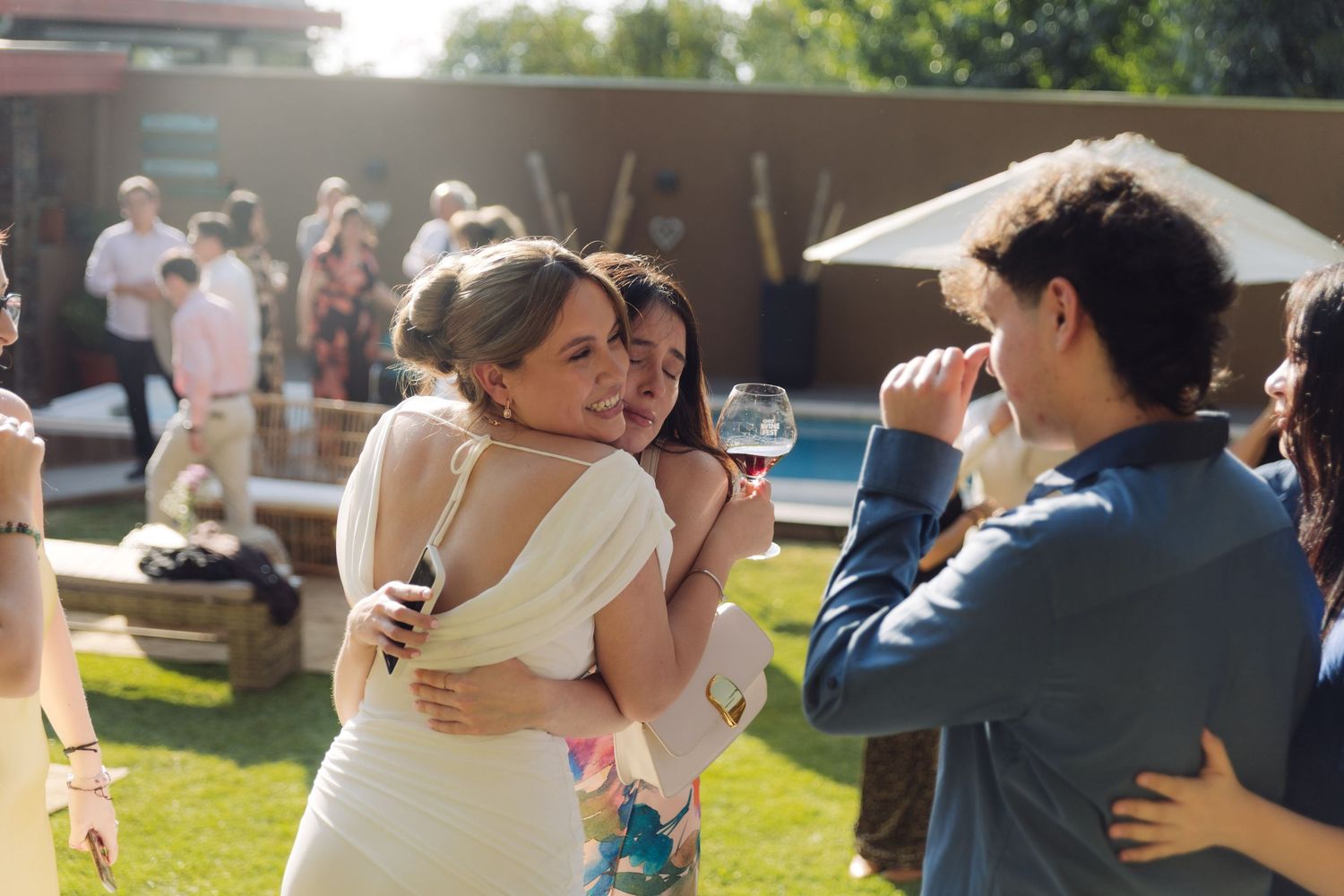 Bride in white dress joyfully hugging a guest near a pool at an outdoor wedding.