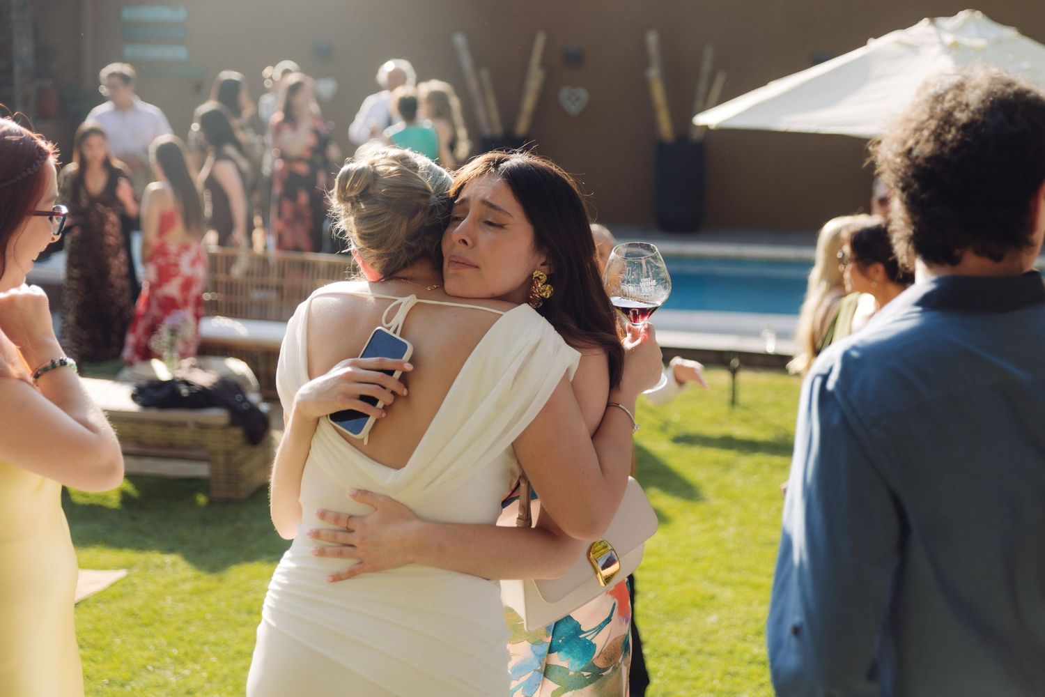Bride in white dress warmly embracing a dark-haired guest at a sunny outdoor wedding.