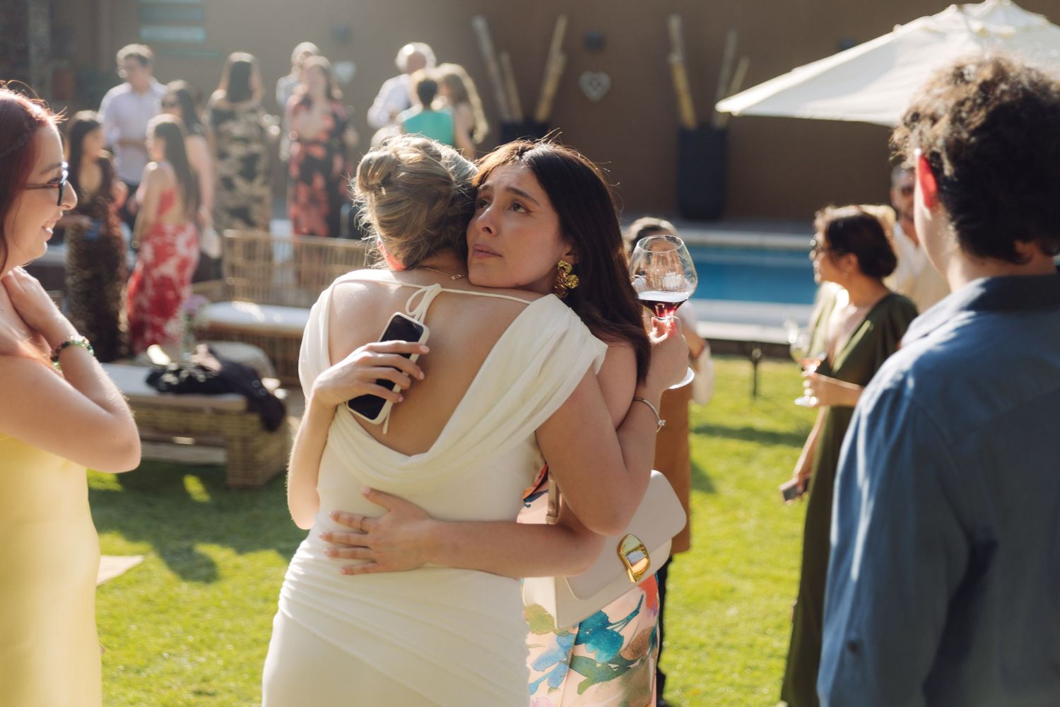 Bride in white dress and dark-haired guest sharing a warm hug at an outdoor wedding.