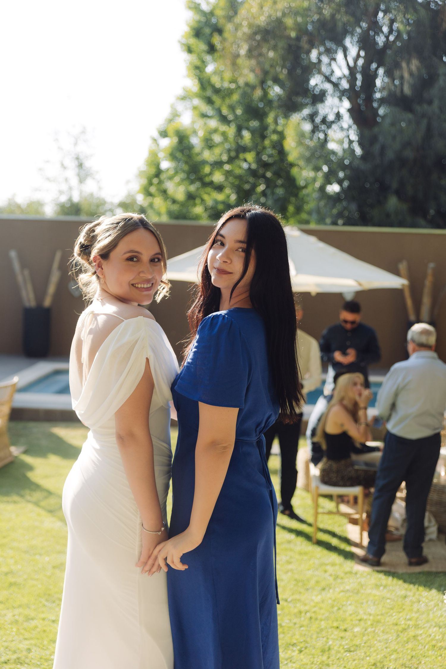 Bride in white gown and bridesmaid in blue dress holding hands at outdoor wedding reception.