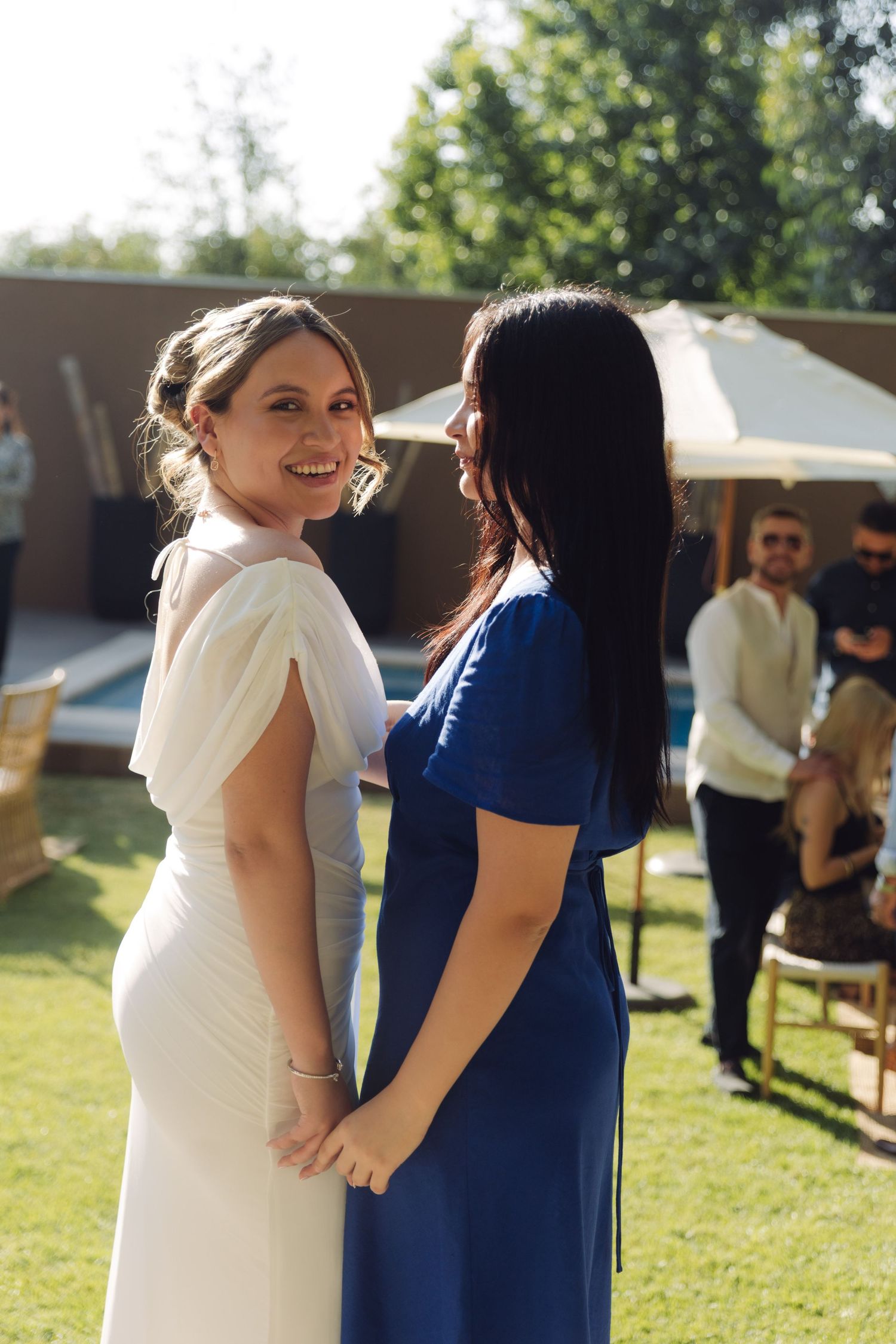 Smiling bride in white dress holding hands with bridesmaid in blue at sunny outdoor wedding.