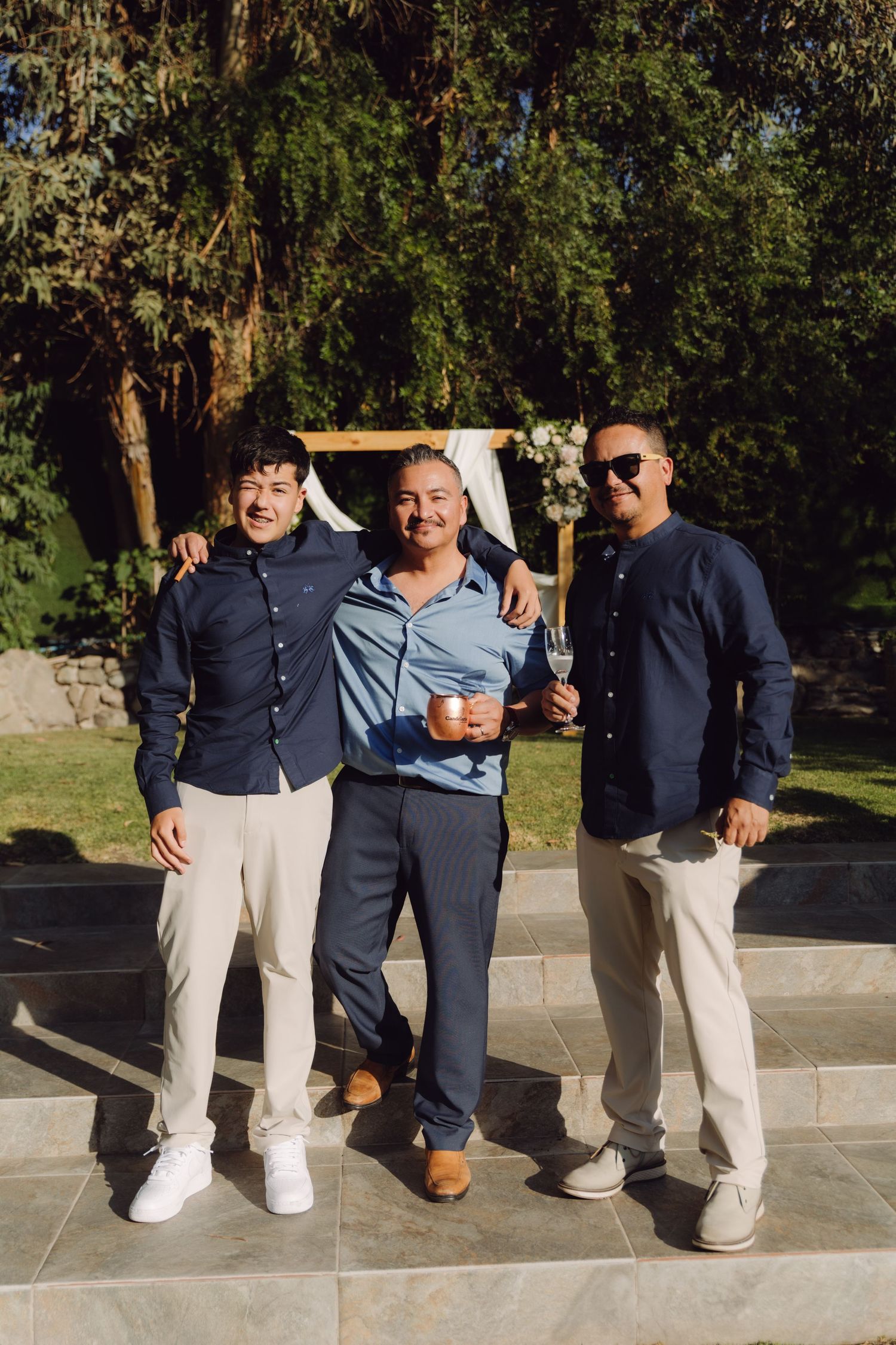 Three men in casual blue shirts smiling together at outdoor wedding ceremony venue.