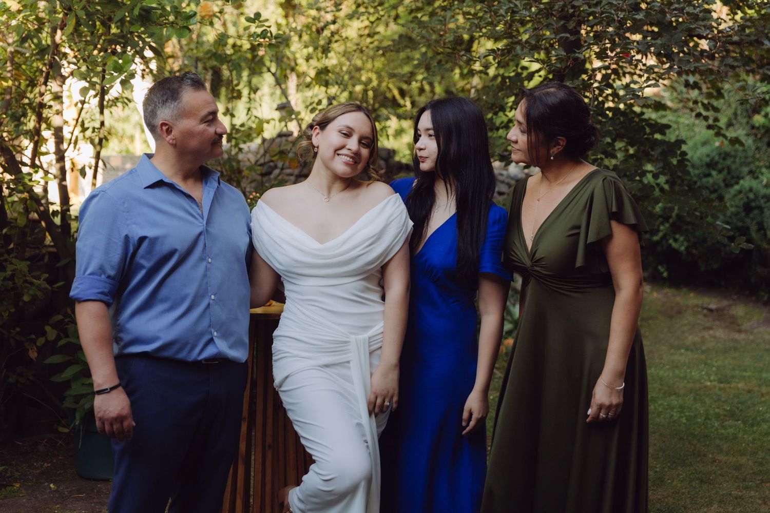 Bride in white gown laughs joyfully with family members in blue and green dresses at outdoor wedding.