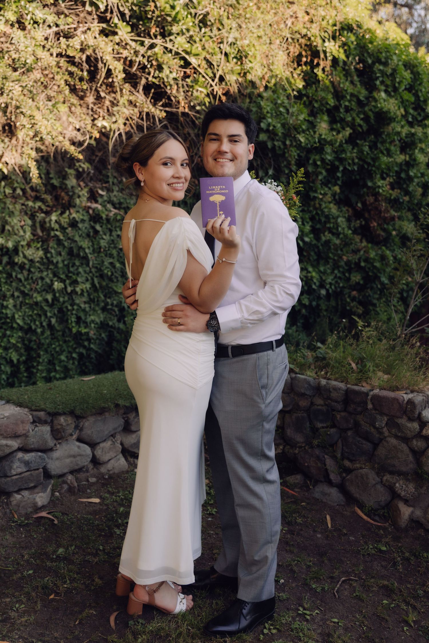Bride and groom posing in garden setting, holding purple book, bride in elegant backless white dress.