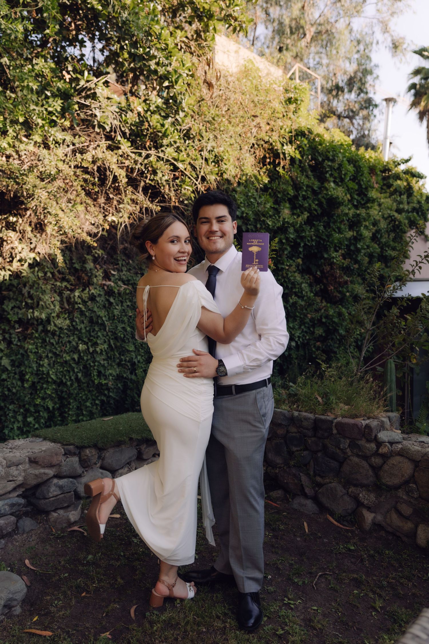 Happy bride and groom embracing in garden, groom holding purple book, bride in backless white dress.