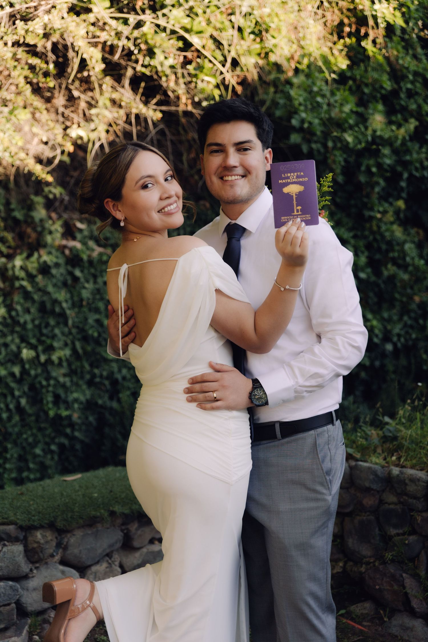 Smiling bride and groom pose together outdoors holding a purple marriage certificate booklet.