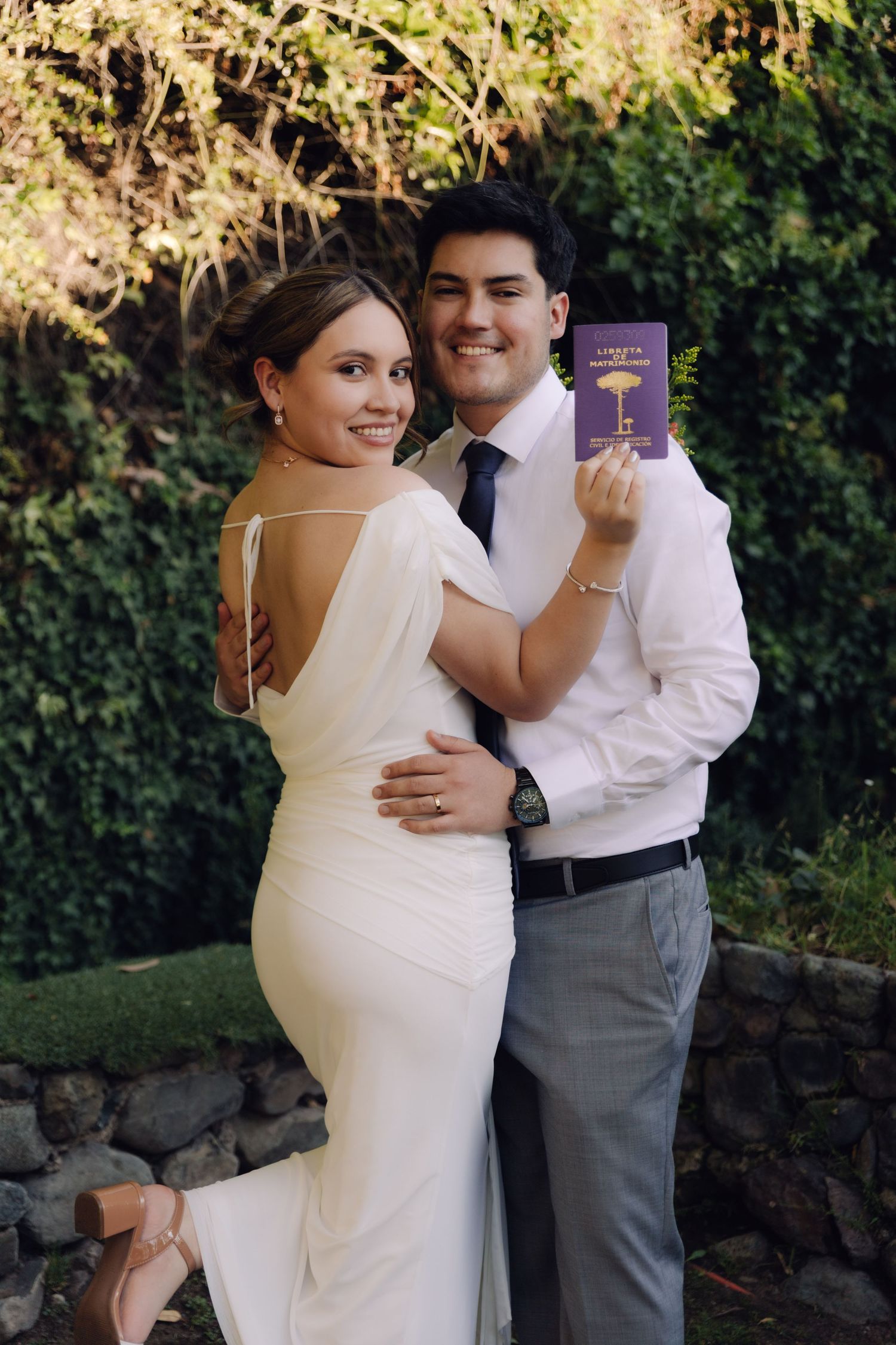 Happy newlyweds embrace outdoors as groom holds purple marriage certificate, bride in backless white gown.