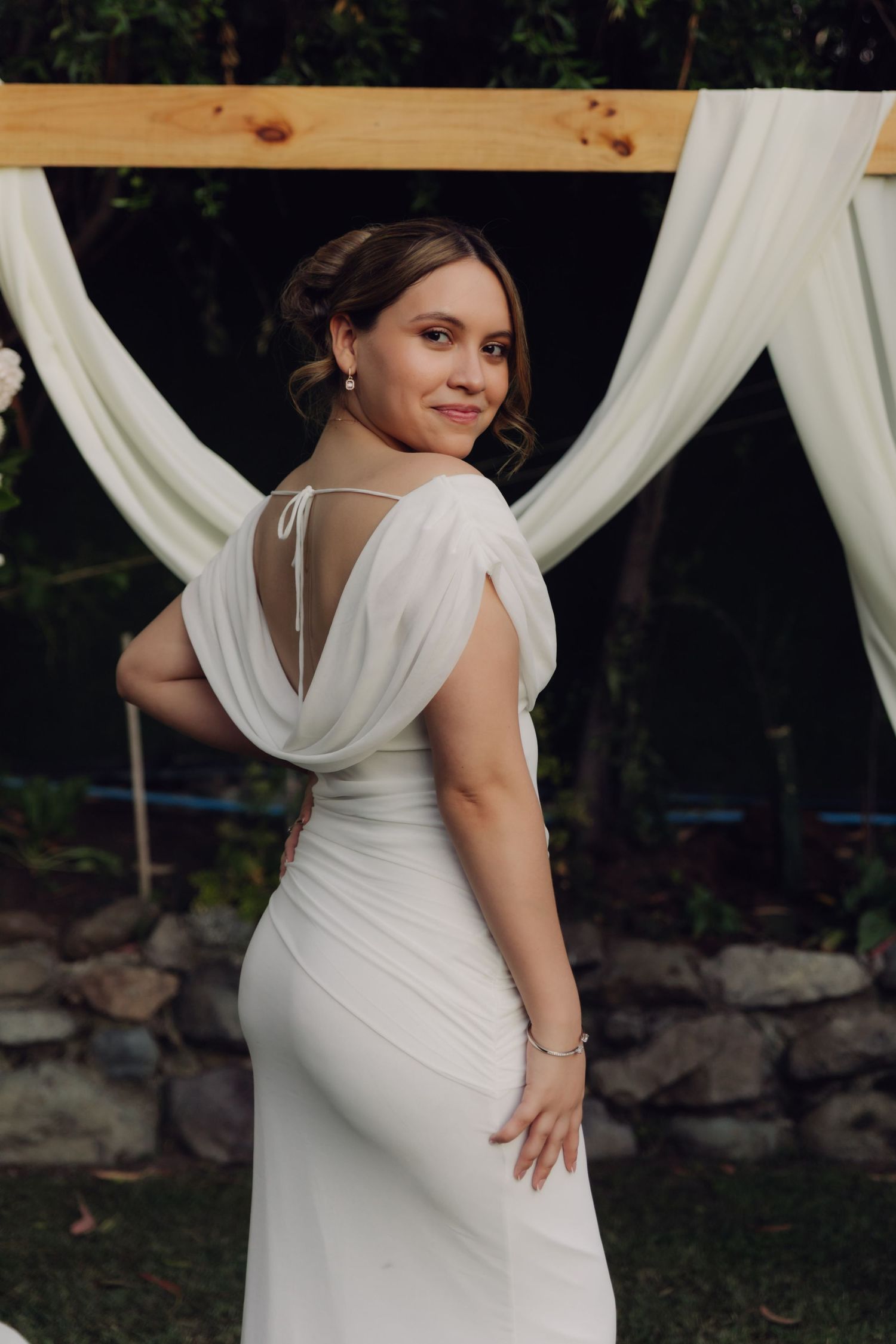 Elegant bride in backless white wedding dress posing in front of draped wooden ceremony arch.