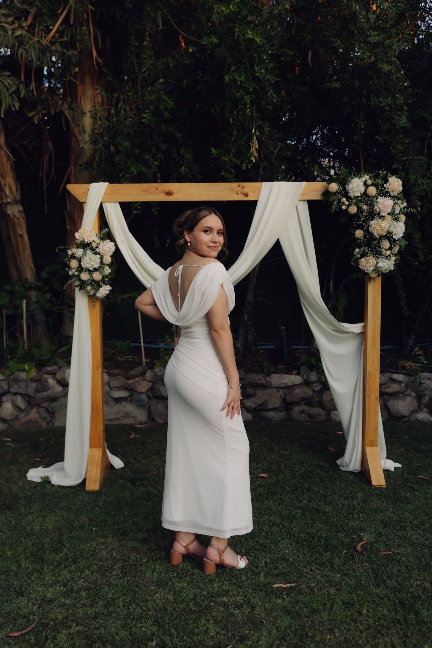 Bride in elegant backless white dress posing in front of wooden arch with white draping and flowers.