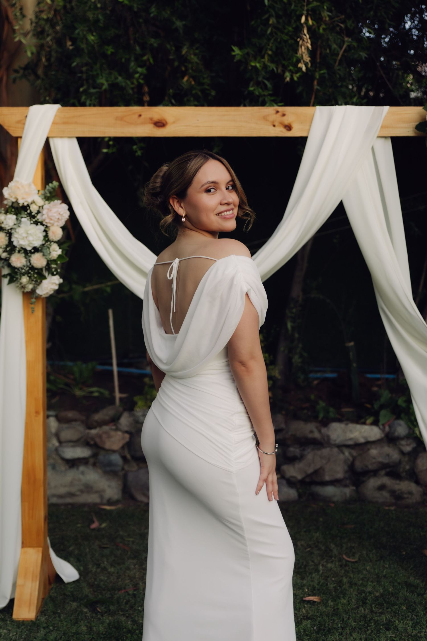 Smiling bride in backless white wedding gown posing before wooden arch with white draping at dusk.