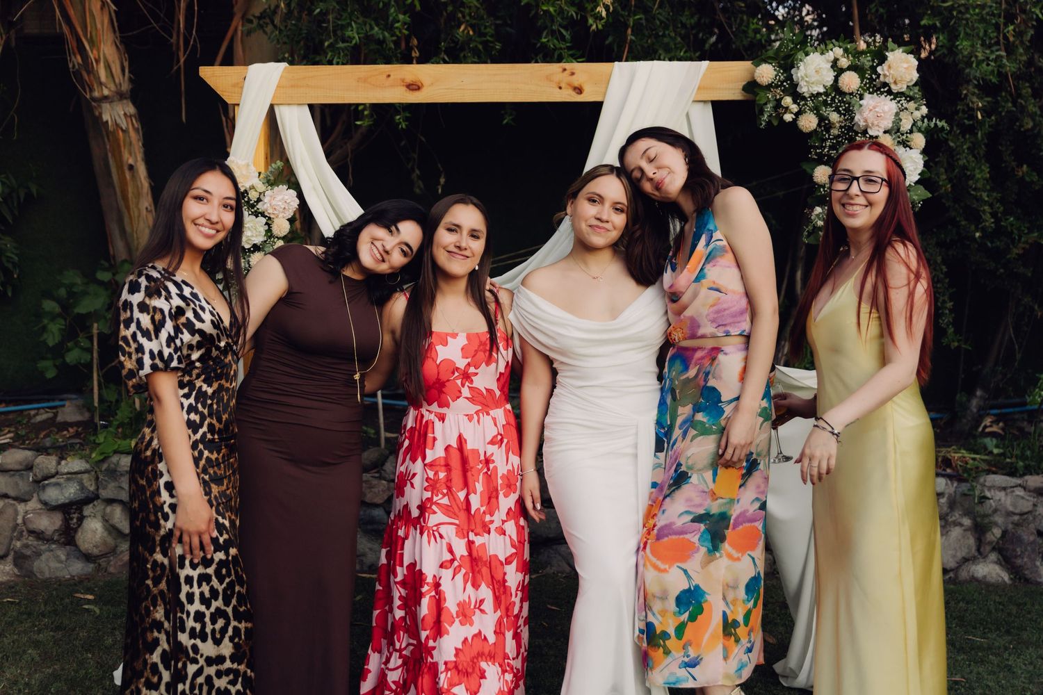 Bride in white off-shoulder gown poses with five female wedding guests in colorful dresses at floral arch.