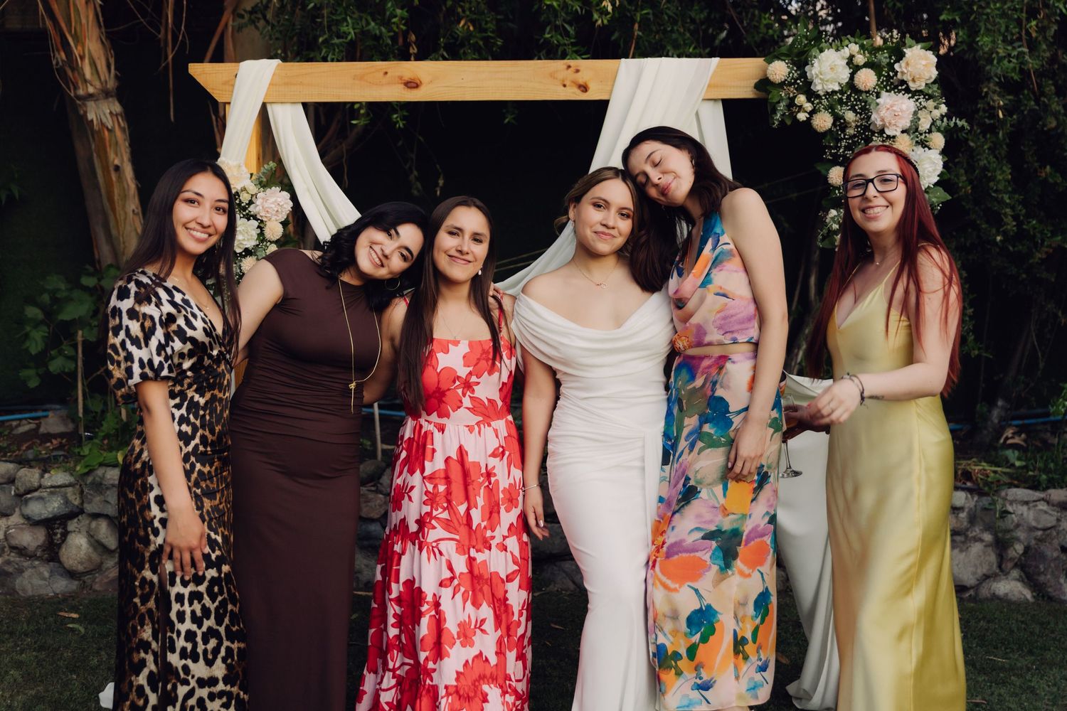 Six women including bride in white gown smile together in front of wooden floral wedding arch.