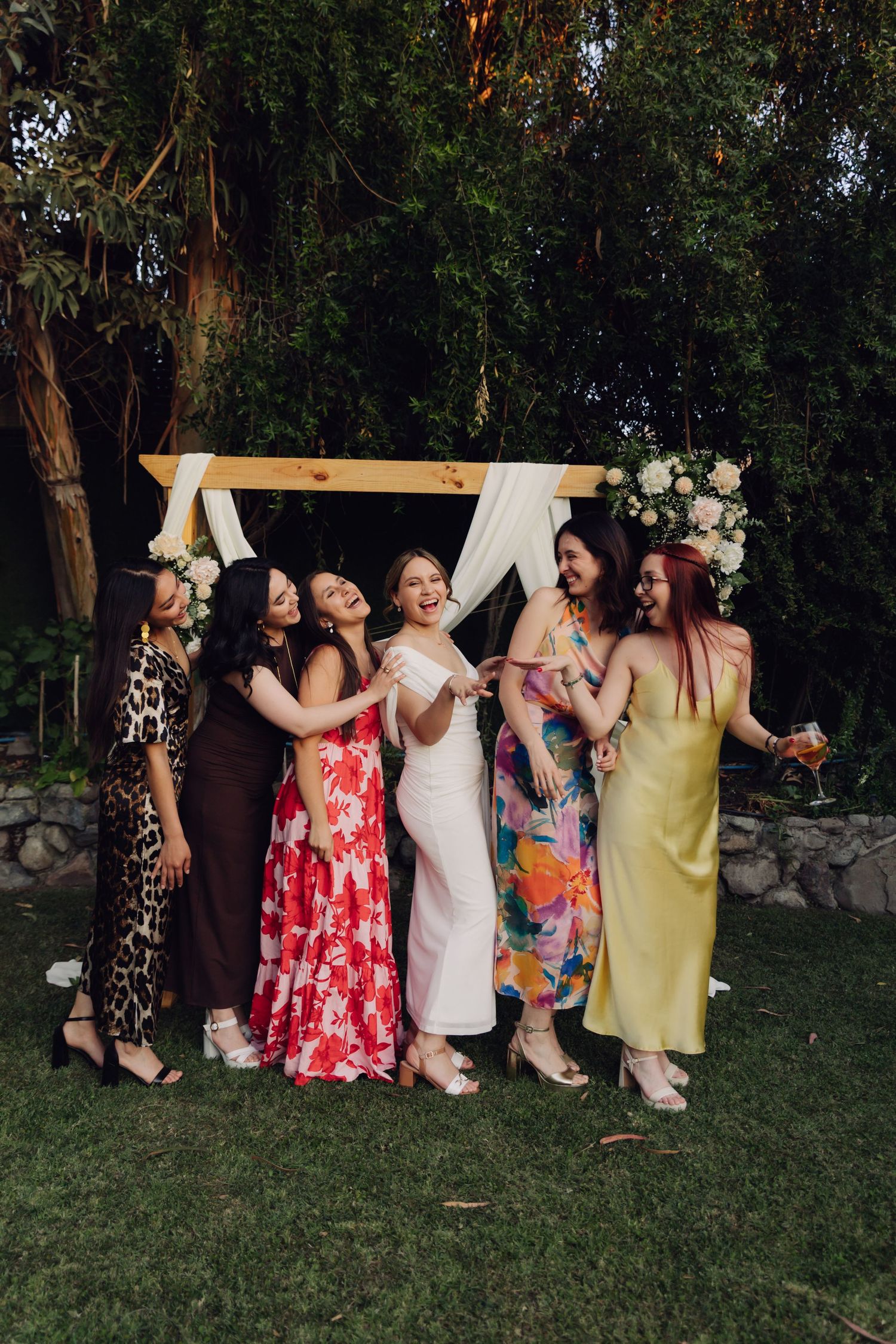 Bride in white dress laughs joyfully surrounded by five friends in colorful gowns at outdoor wedding.