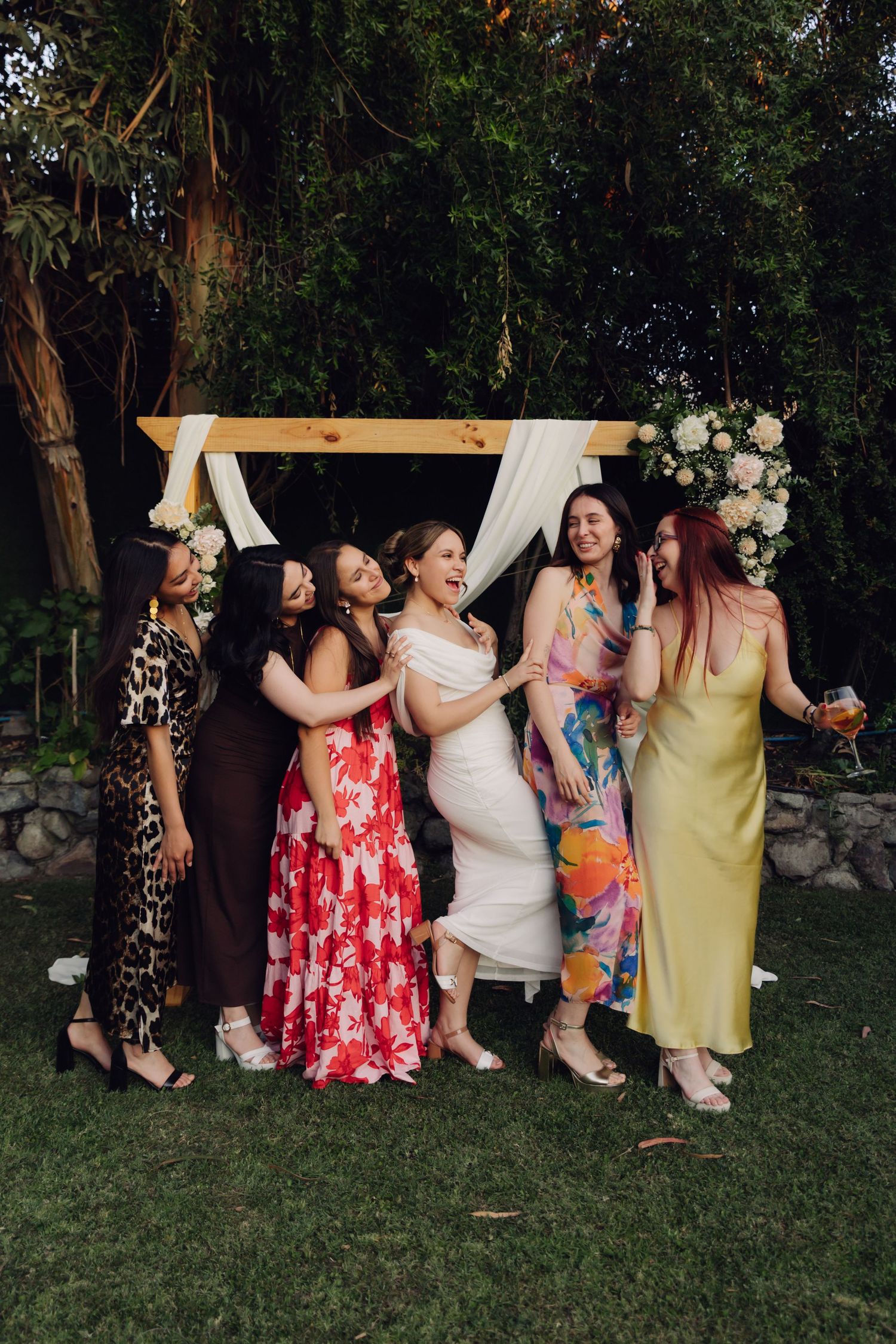 Bride and five guests in vibrant dresses laugh and celebrate together beneath decorated outdoor wedding arch.