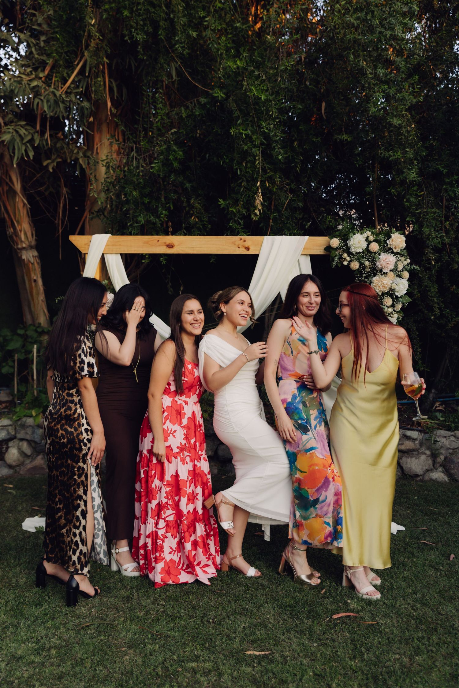 Six women in elegant dresses pose and laugh together at an outdoor wedding ceremony under a wooden arch.