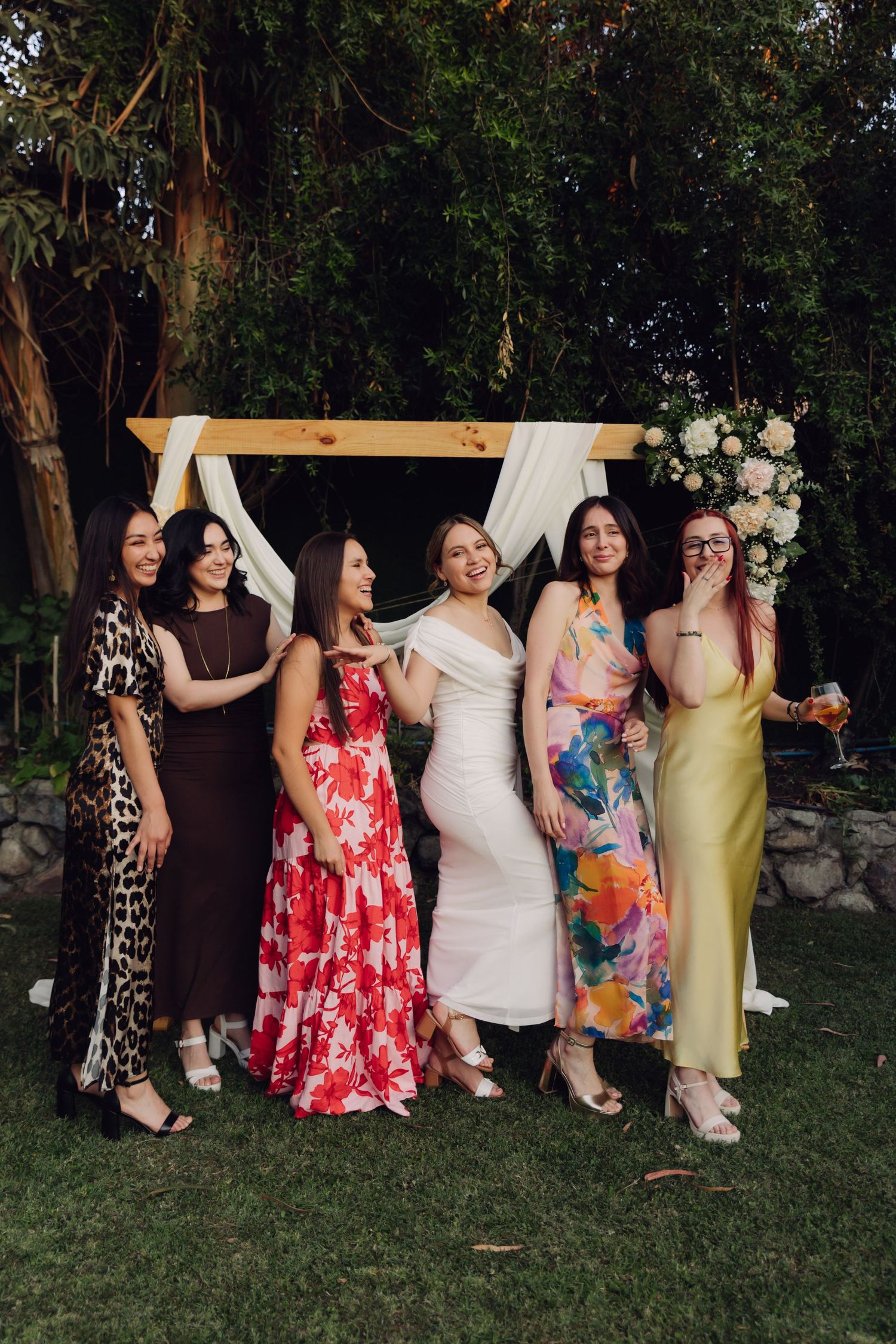 Group of six women in colorful formal dresses smiling together at an outdoor garden wedding celebration.