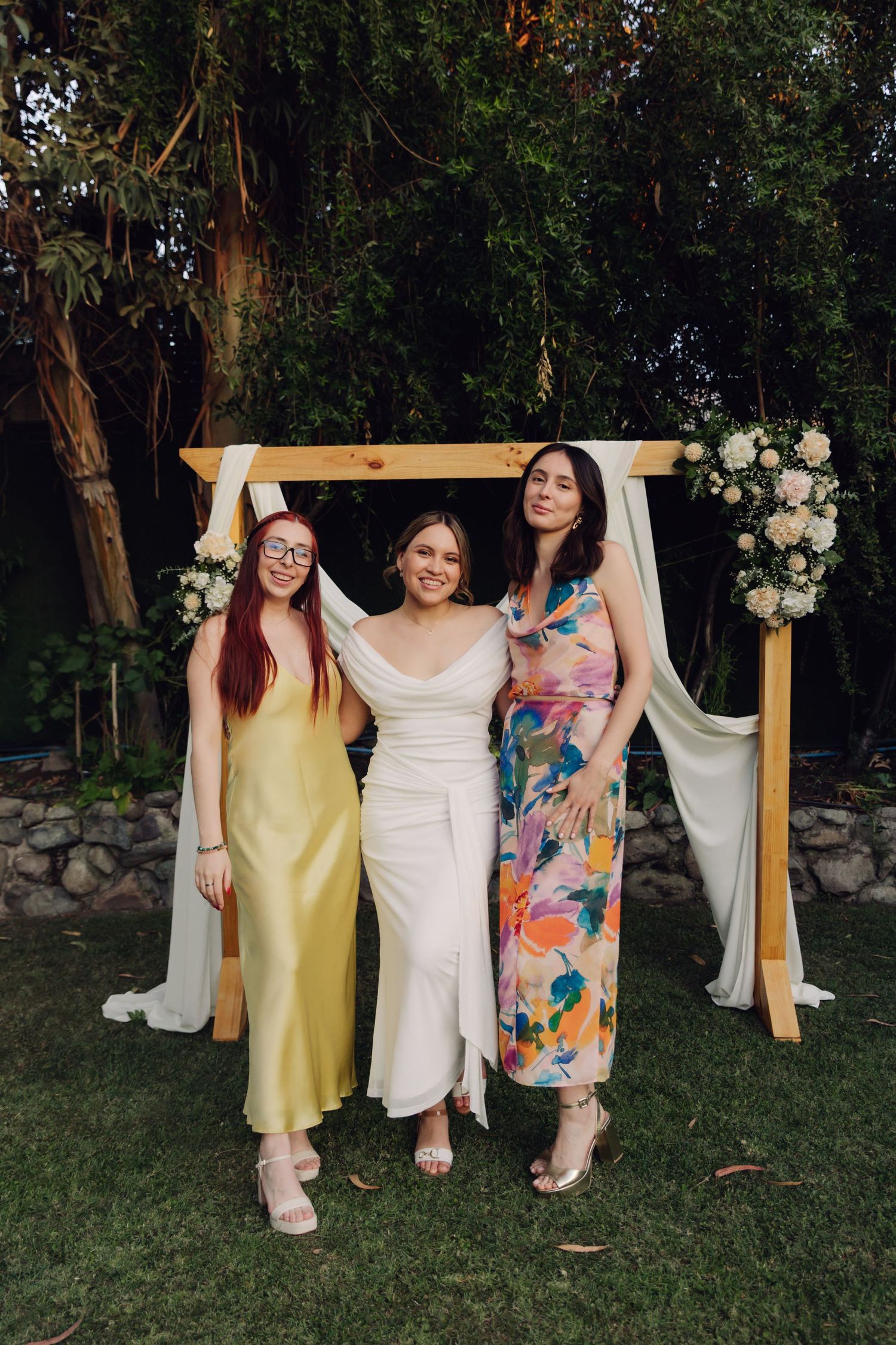 Bride in white dress poses with two friends in yellow and floral dresses at decorated wedding arch.