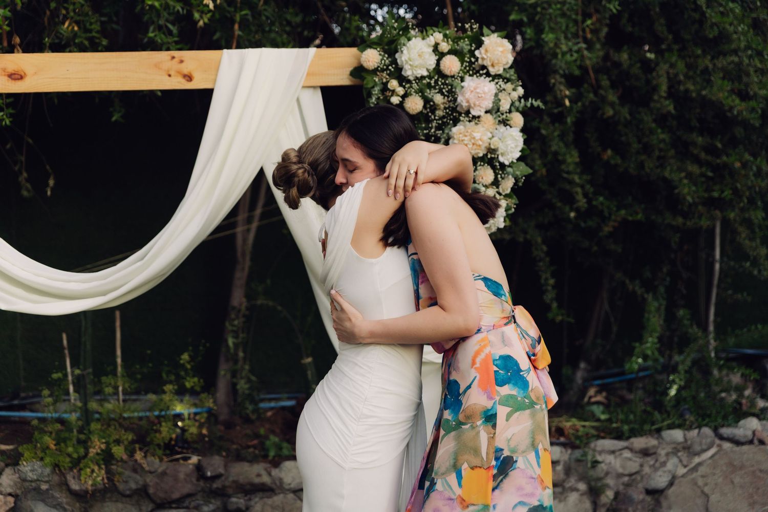Bride in white wedding dress shares a warm embrace with a guest in a colorful floral dress.