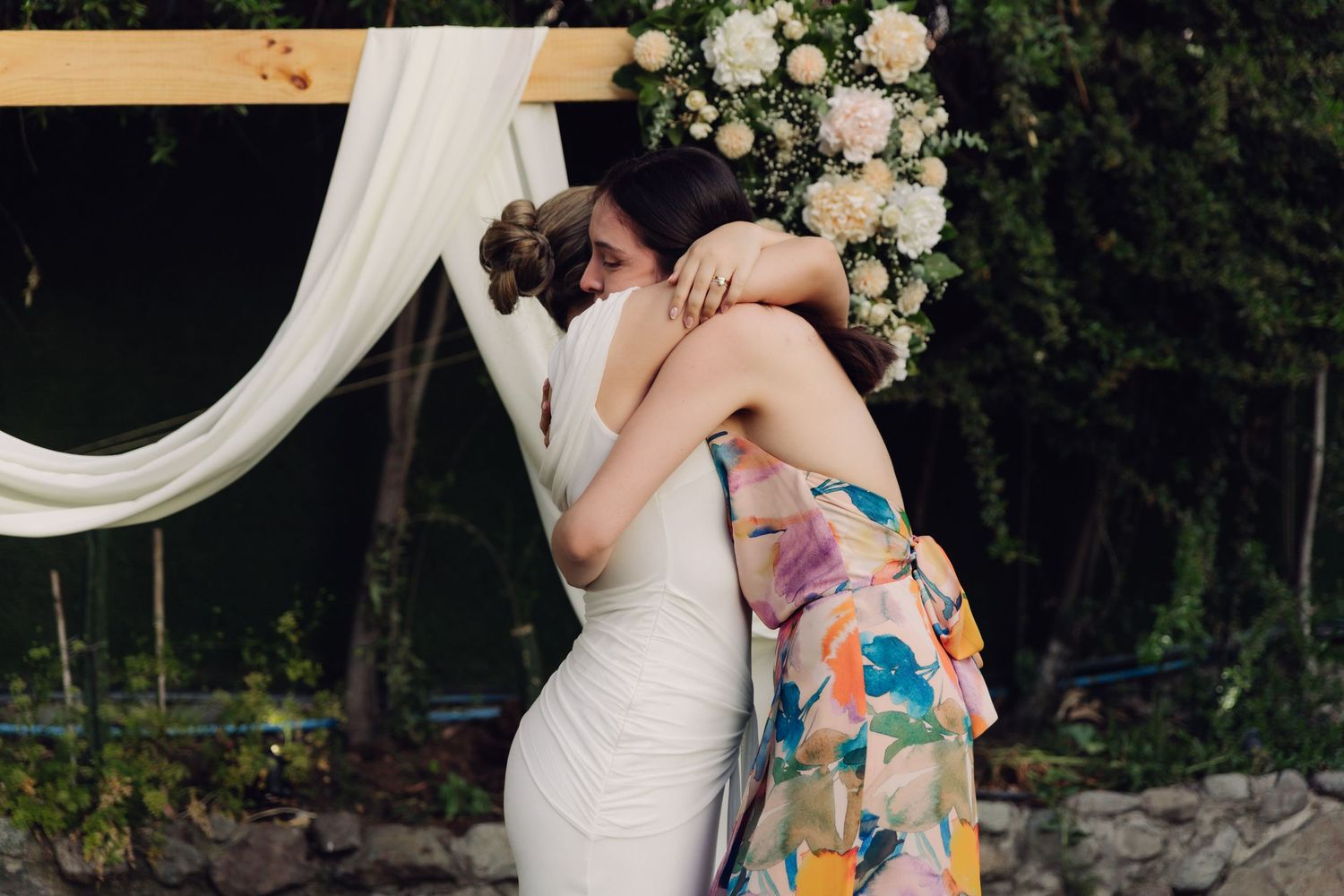 Two women share an emotional hug at an outdoor wedding near a wooden arch with floral arrangements.