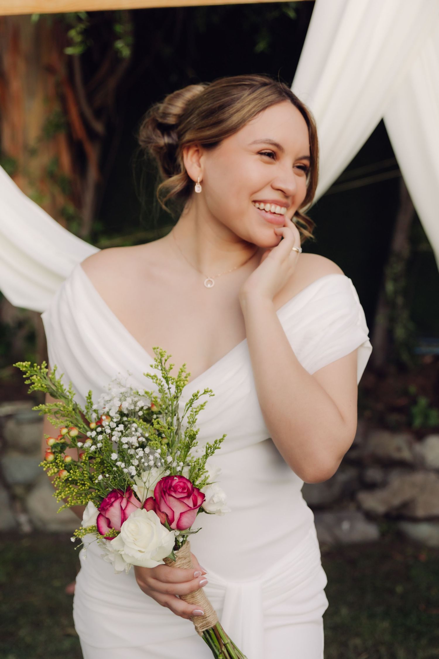 Radiant bride in white dress laughing while holding colorful bouquet under draped wedding ceremony arch.